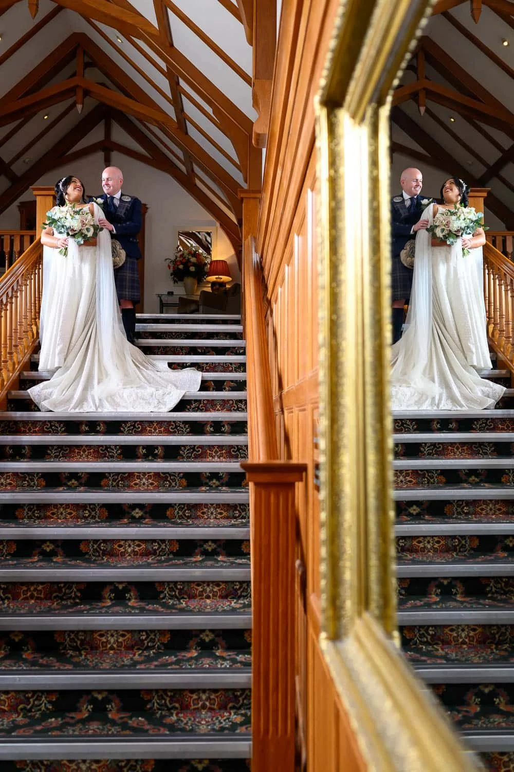 A relaxed and joyful wedding moment at Lochgreen House Hotel in Troon, Ayrshire, as the bride and groom share a smile at the top of the grand staircase. Captured by a Lochgreen House Hotel wedding photographer, this image uses a mirror reflection to 