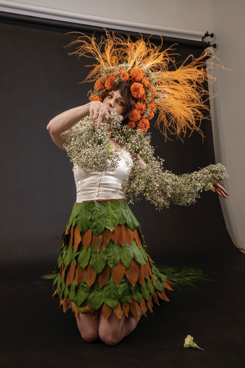 A woman kneels on the floor wearing a costume made of floral and leaf elements, with a large floral headdress featuring orange flowers and feathers.