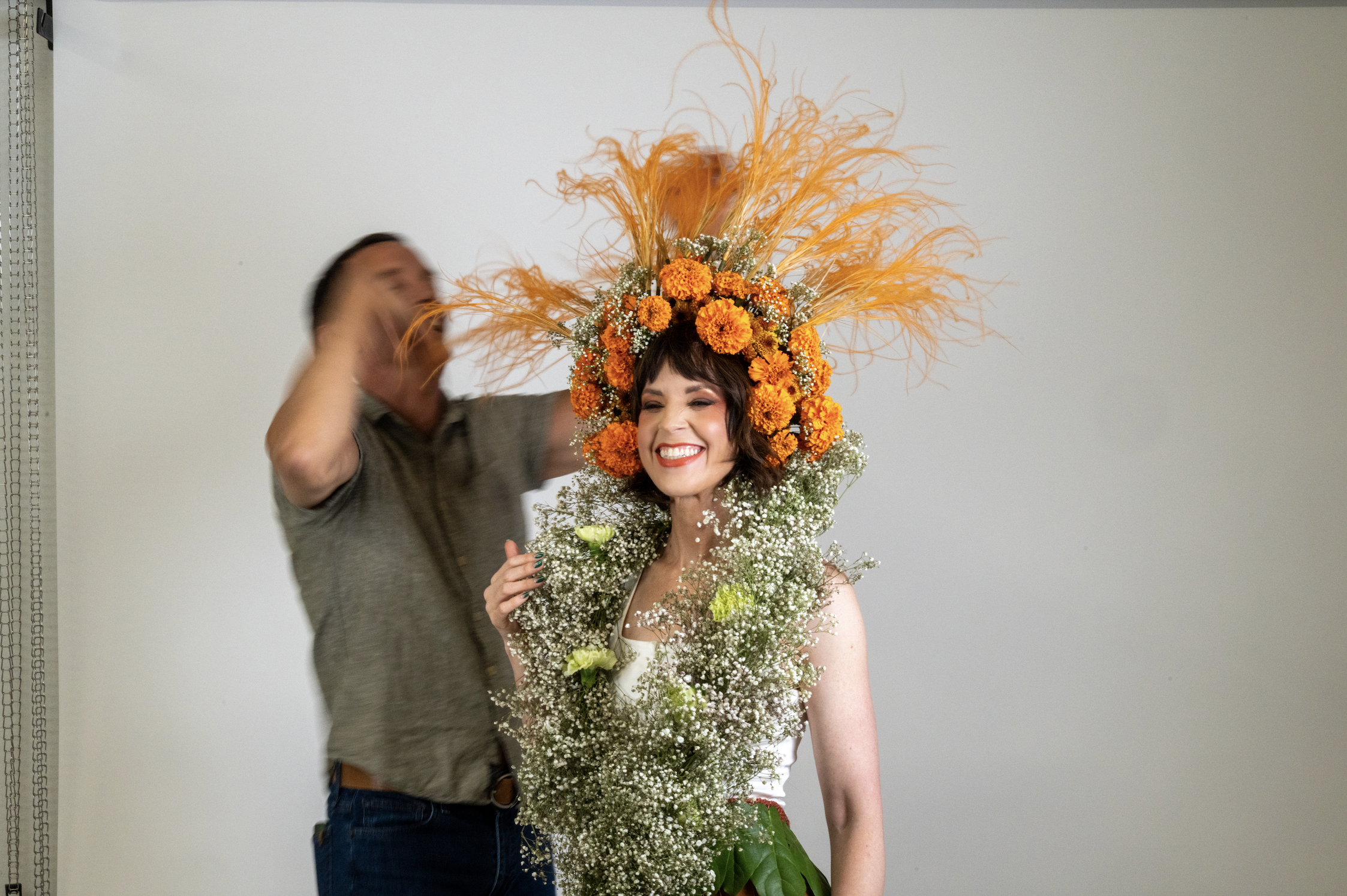 A woman with a big smile and wearing an elaborate floral headpiece made of orange marigolds, baby's breath, and orange feathers. A man behind her is adjusting the headpiece.