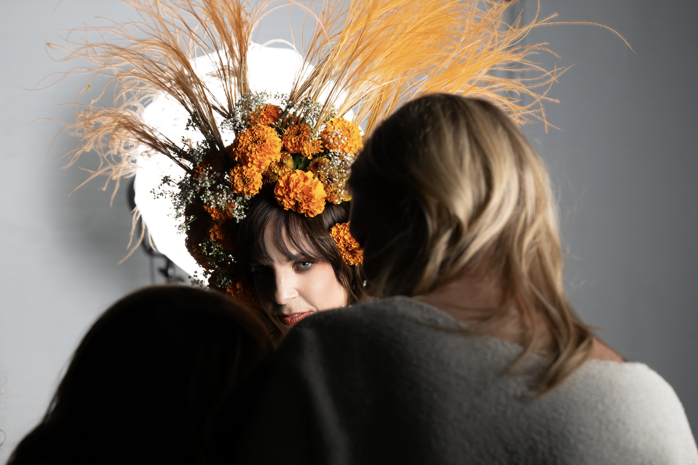 A woman wearing a large floral headpiece with orange marigolds, pampas grass, and small white flowers, being photographed in front of a bright studio light.