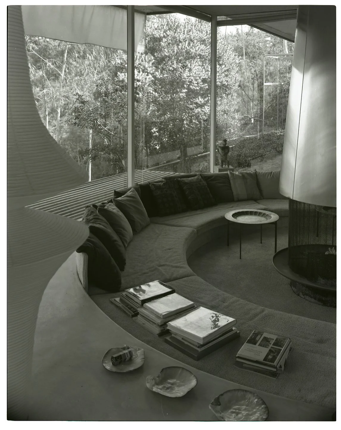 Interior living room with curved sofa, books, and magazines on a table, large windows showing trees outside, and a sculpture of a dog outside.