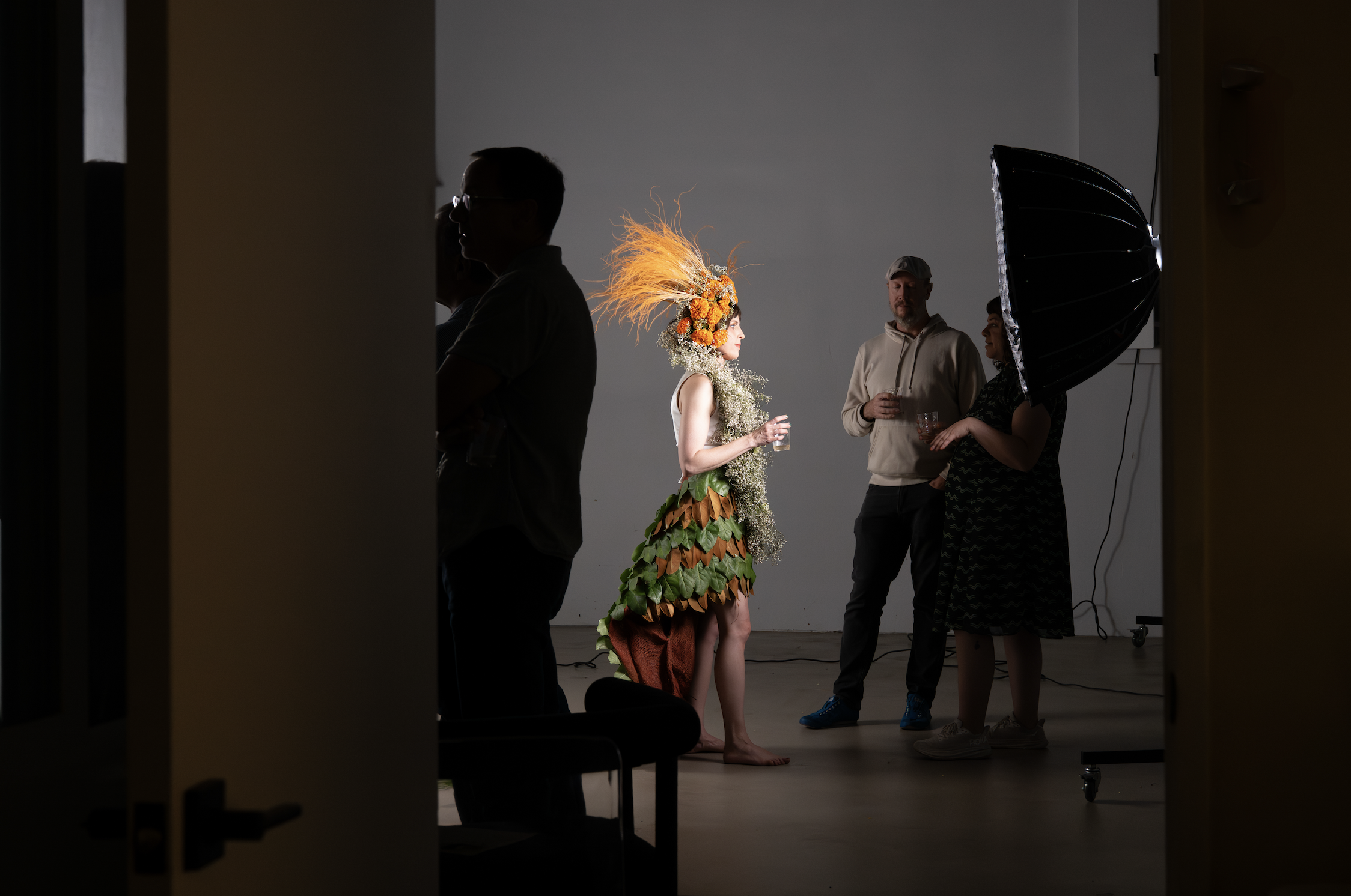 Behind-the-scenes of a photo shoot with a model in a nature-inspired costume and flowers in her hair, being photographed by a professional with studio lighting, while assistants observe.