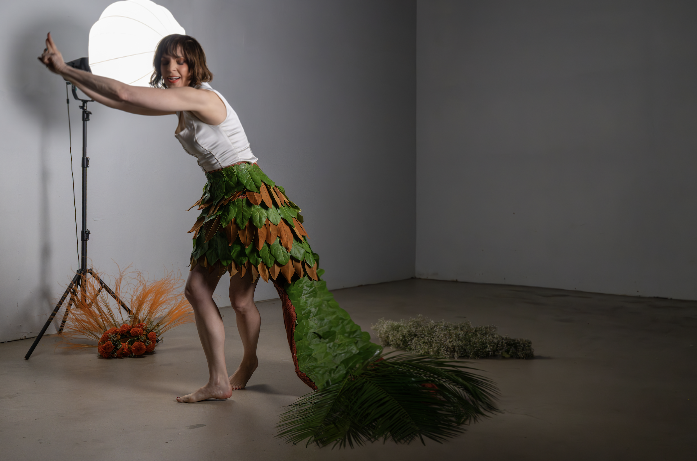 A woman performing on stage wearing a costume with layers of green and brown leaves, a long green tail with brown accents, surrounded by plant props, standing barefoot on a plain floor against a gray background, with a large light on a stand behind h
