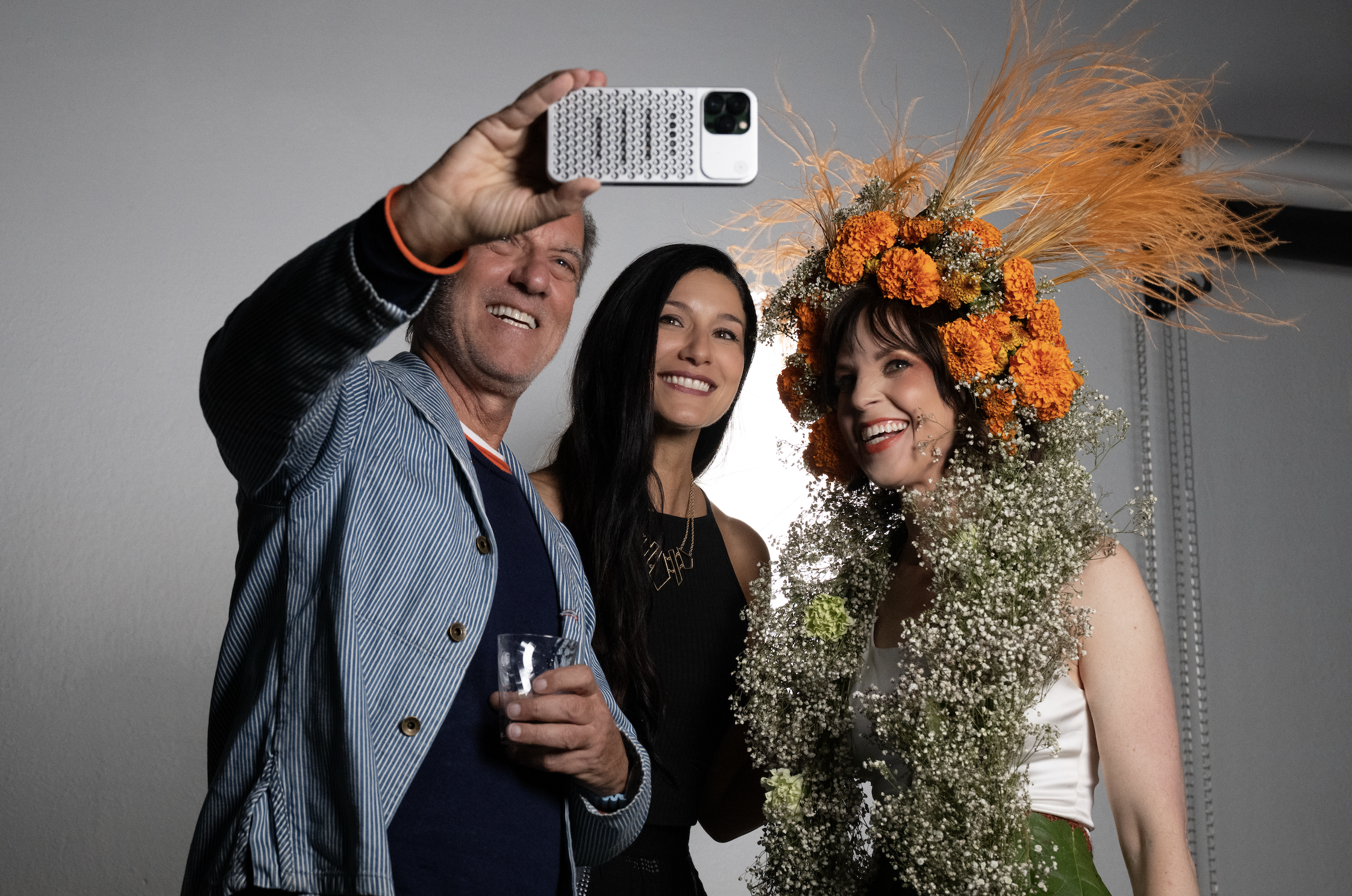 Three people taking a selfie. One woman is wearing an extravagant floral headdress with orange flowers and pampas grass. The group is smiling and holding drinks.