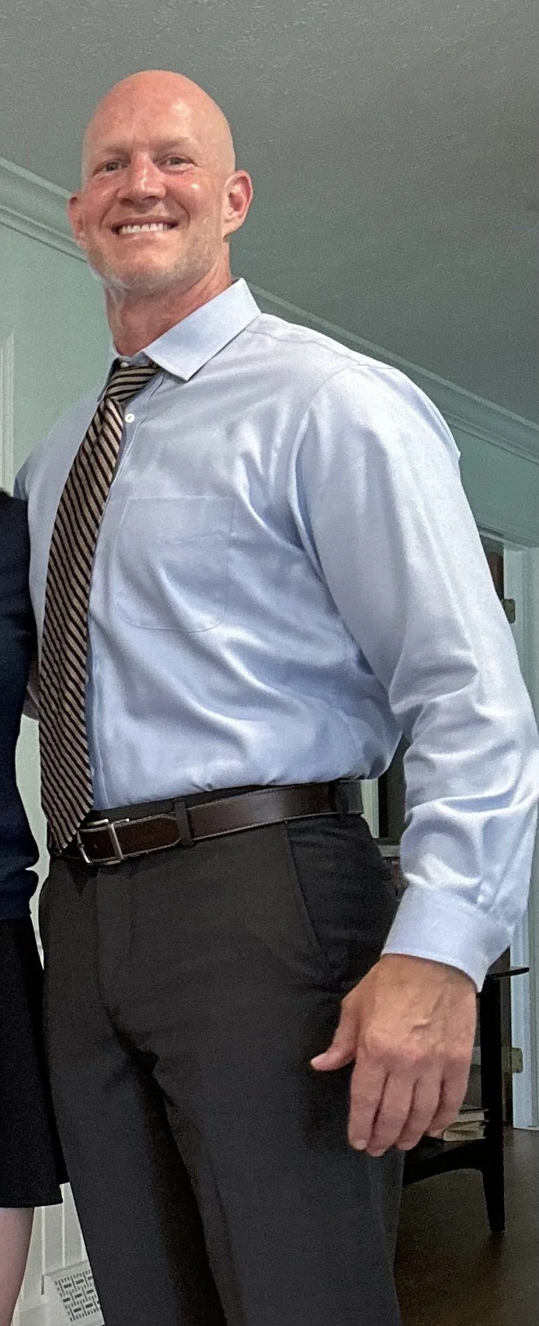 Bald man in a light blue dress shirt and striped tie smiling at the camera, standing indoors.