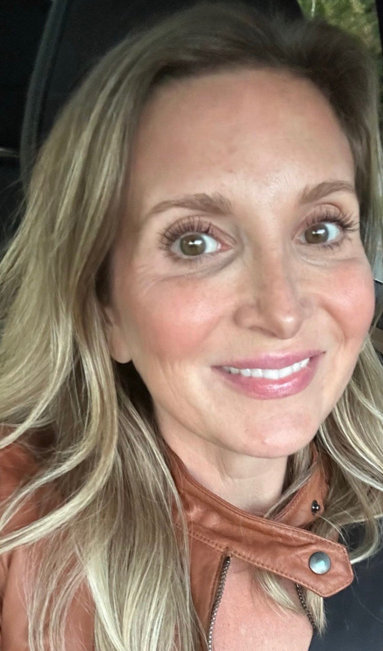 Close-up of a woman with long blonde hair, wearing a brown leather jacket, smiling, with a blurred background.