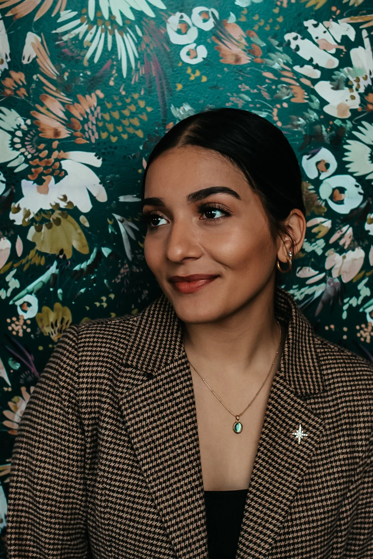 A woman with dark hair styled back, wearing a houndstooth blazer, gold hoop earrings, and layered gold necklaces with a green pendant, posing in front of a vibrant floral-patterned background.