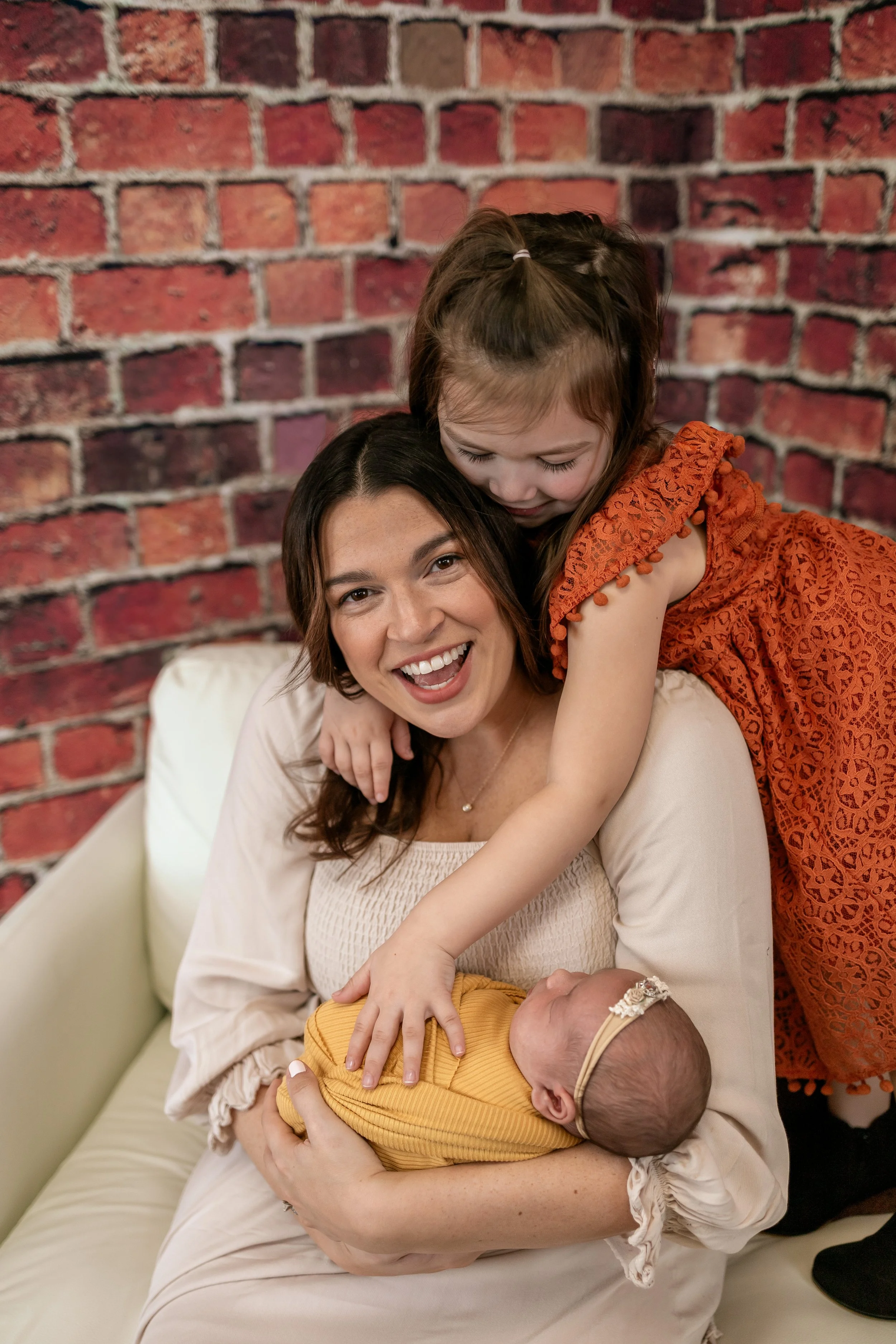 A woman holding a sleeping baby in her arms, a young girl in an orange dress leaning over her, hugging her neck, and smiling happily in front of a red brick wall.