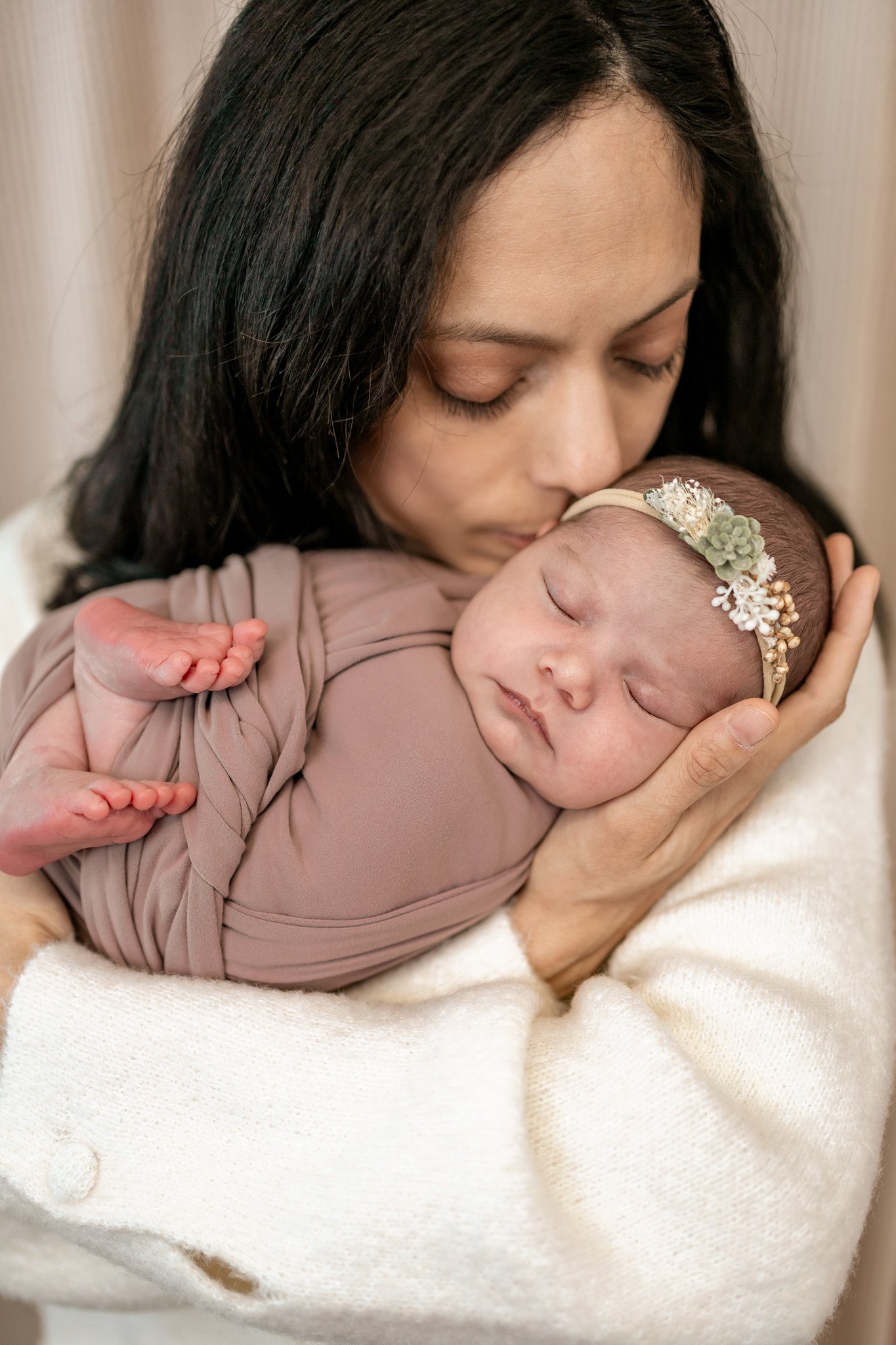 A woman gently holding a sleeping newborn baby close to her face, with the woman kissing the baby's forehead. The baby is wearing a floral headband and wrapped in a mauve cloth, while the woman is dressed in a cream sweater.