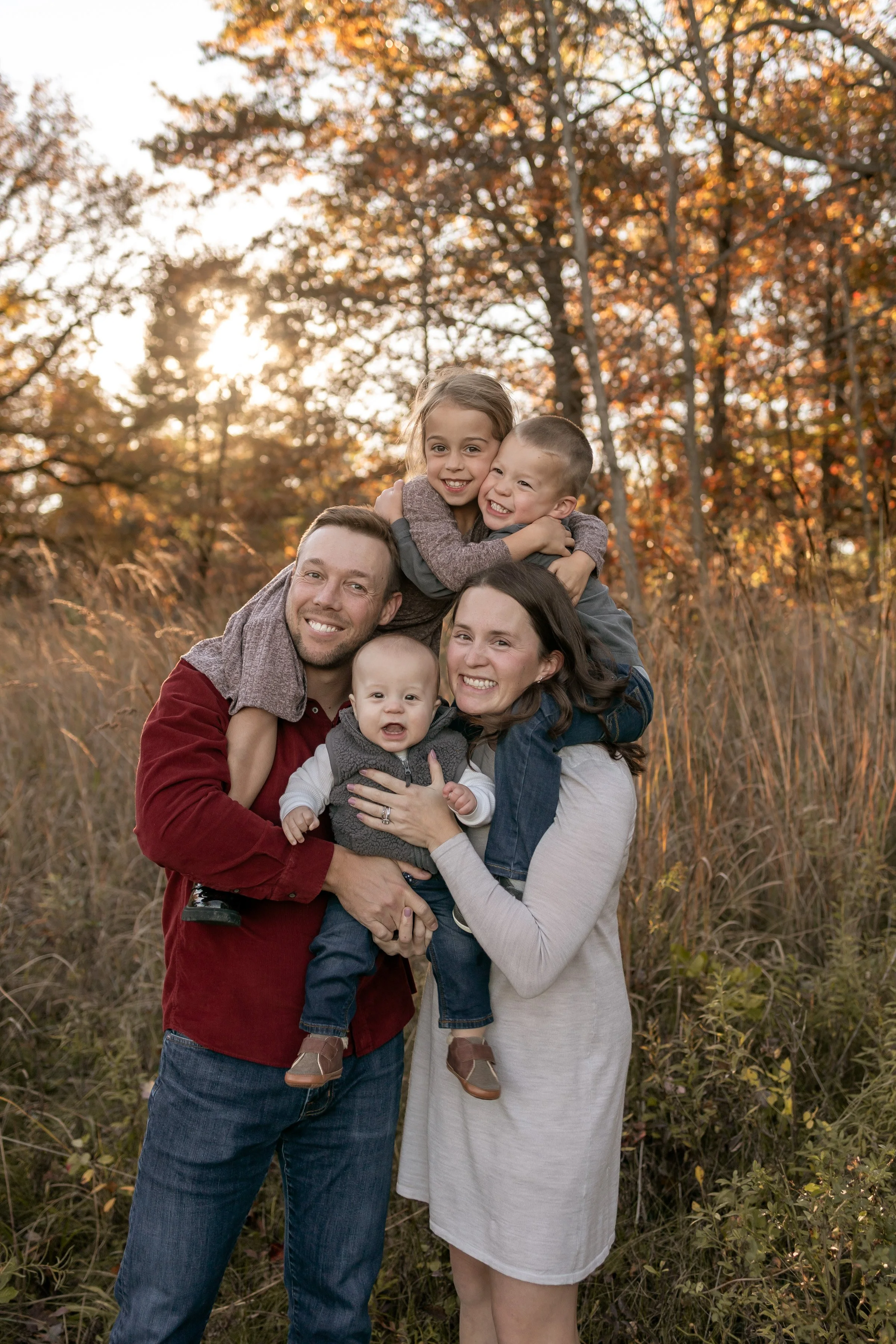 Family of five outdoors in a field of tall grass during fall, smiling and hugging each other with autumn trees and sunlight in the background.