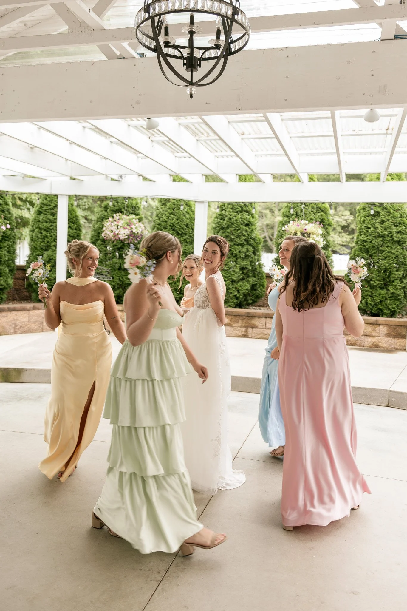 Bridesmaids dressed in colorful dresses are dancing and celebrating at an outdoor wedding reception under a white pavilion with hanging flowers and greenery in the background, during the daytime.
