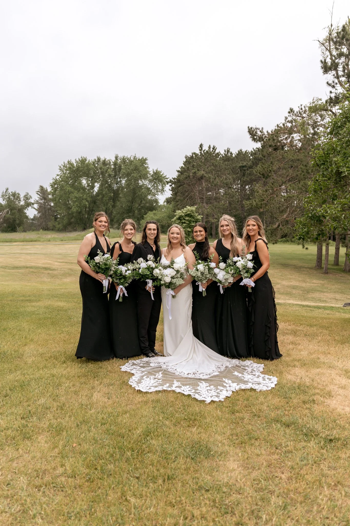 Group of seven women, including a bride in a white wedding dress and six bridesmaids in black dresses, posing outdoors on a grassy field with trees in the background, under an overcast sky, holding bouquets of white and green flowers.