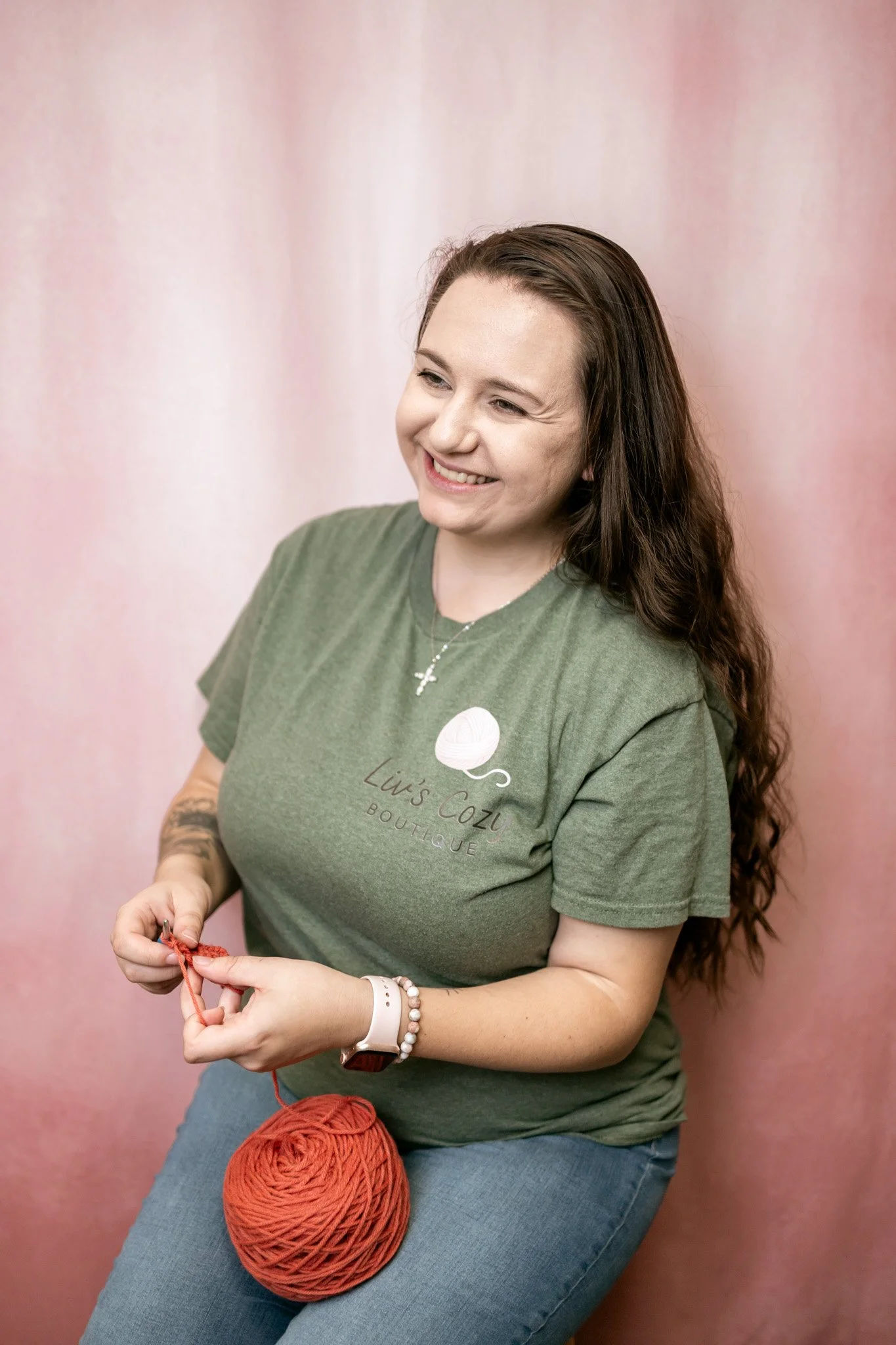 A woman with long brown hair, wearing a green t-shirt with 'Liv's Cozi Boutique' and a knitting ball logo, smiling while holding red yarn and knitting needles.