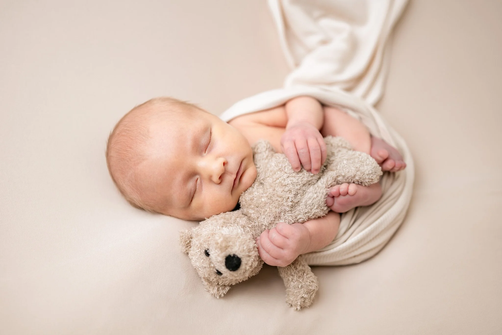 A sleeping newborn baby lying on a soft surface, holding a small plush teddy bear, wrapped in a light-colored blanket.
