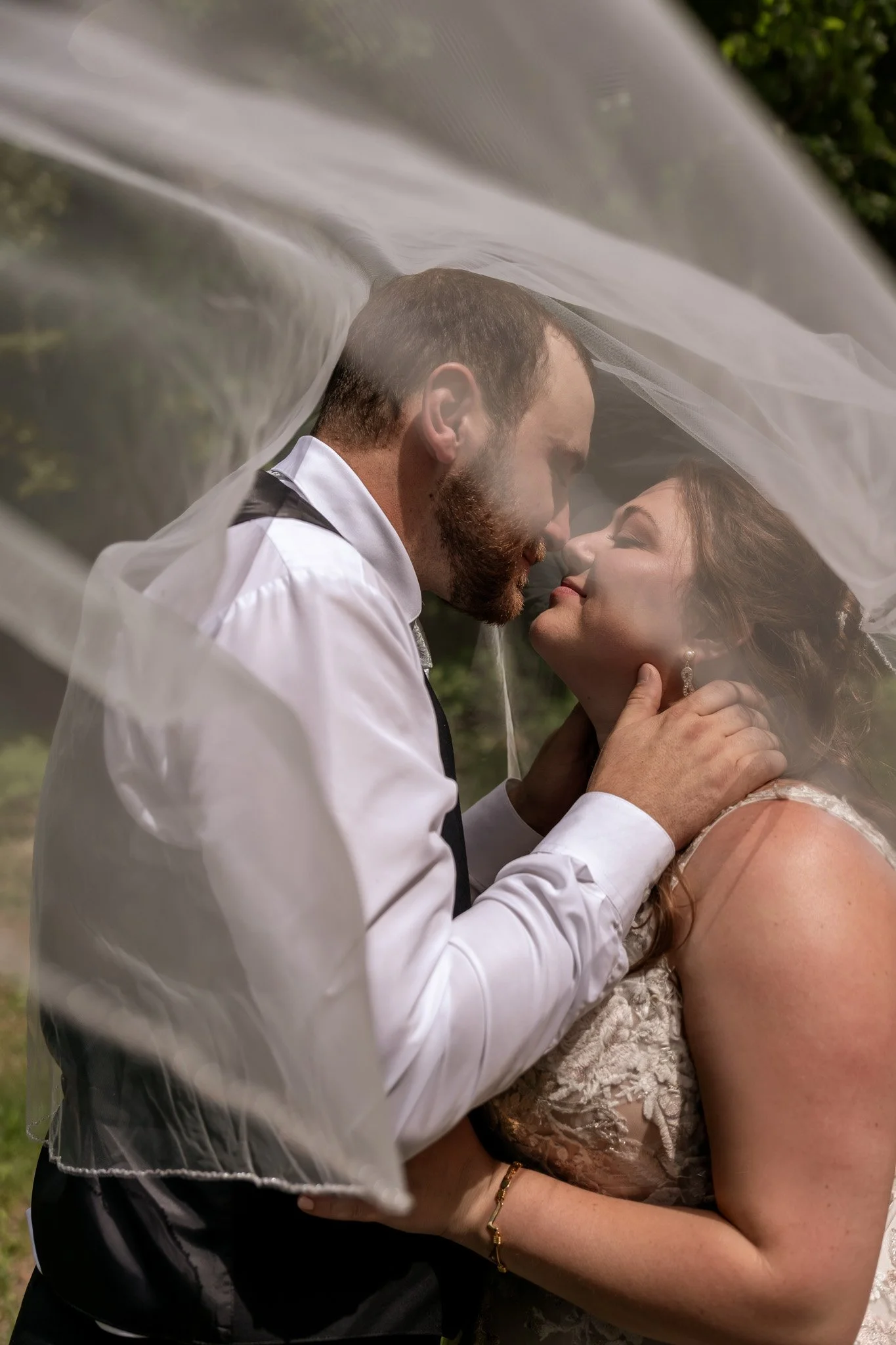 A man and woman are kissing under a sheer white veil, with the man wearing a white shirt and vest, and the woman an elegant lace dress with jewelry.