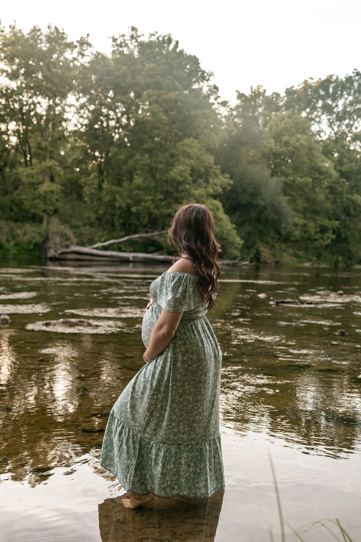 A pregnant woman with long, wavy brown hair standing on a rock in a river, wearing a green floral dress, looking away from the camera, with a background of trees and a partly cloudy sky.