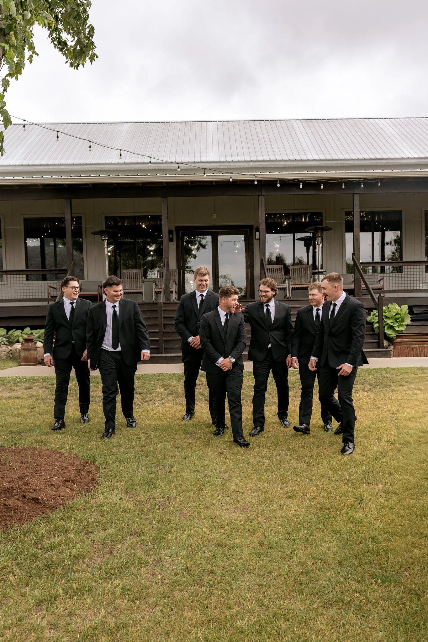A group of seven men in suits walking and talking on a grassy lawn outside a building with large windows, a deck, and string lights overhead.