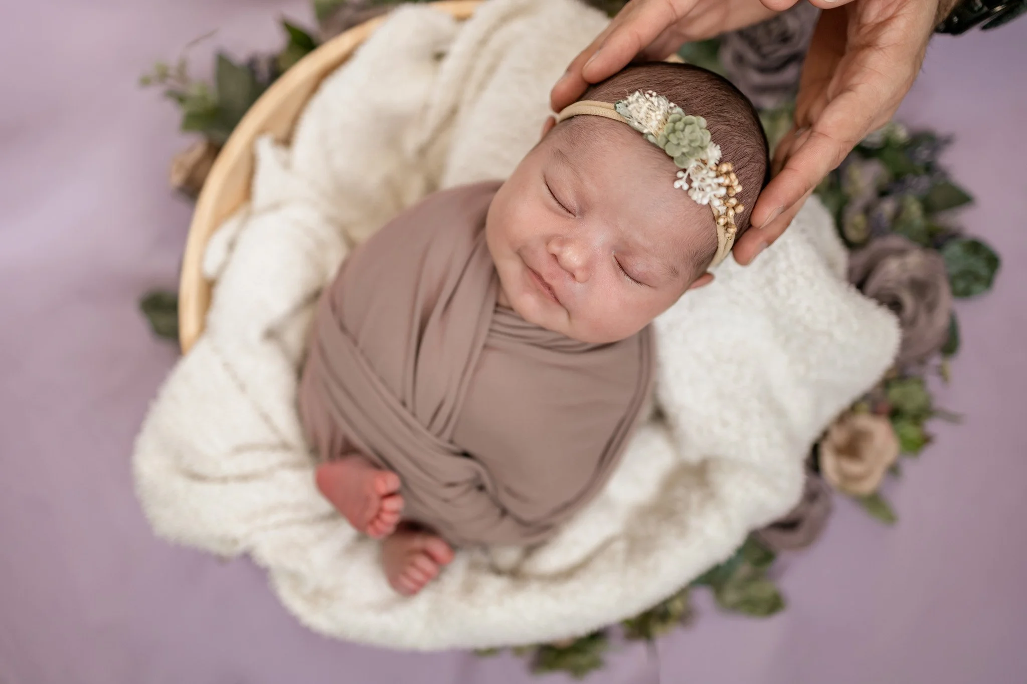A sleeping baby wrapped in a taupe blanket, lying on a white fluffy blanket in a basket, with a floral headband, being gently touched on the head by an adult hand, surrounded by purple flowers.