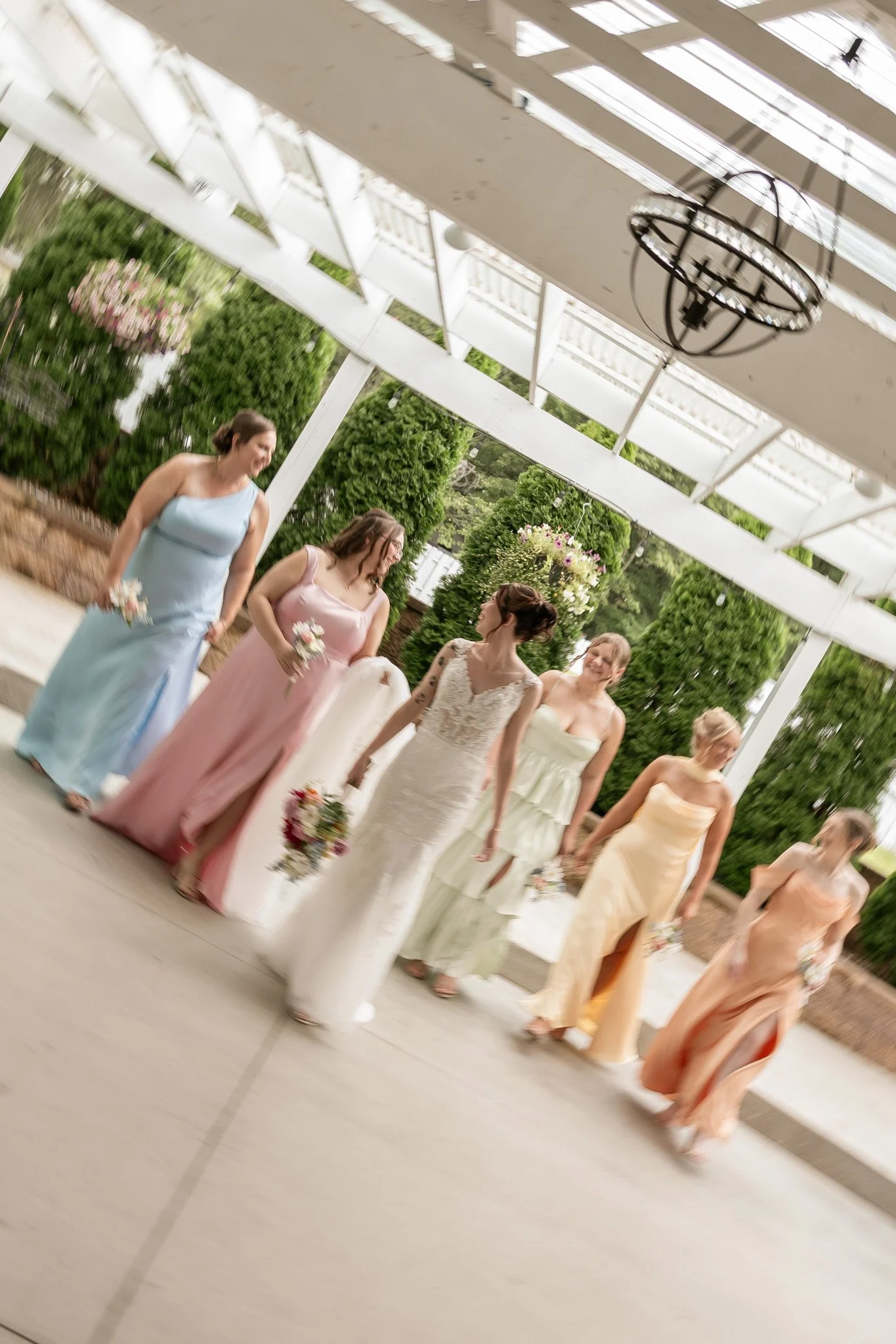 Bridesmaids in colorful dresses standing in a line inside a modern, glass-roofed building with hanging light fixtures and green outdoor scenery visible through the windows.