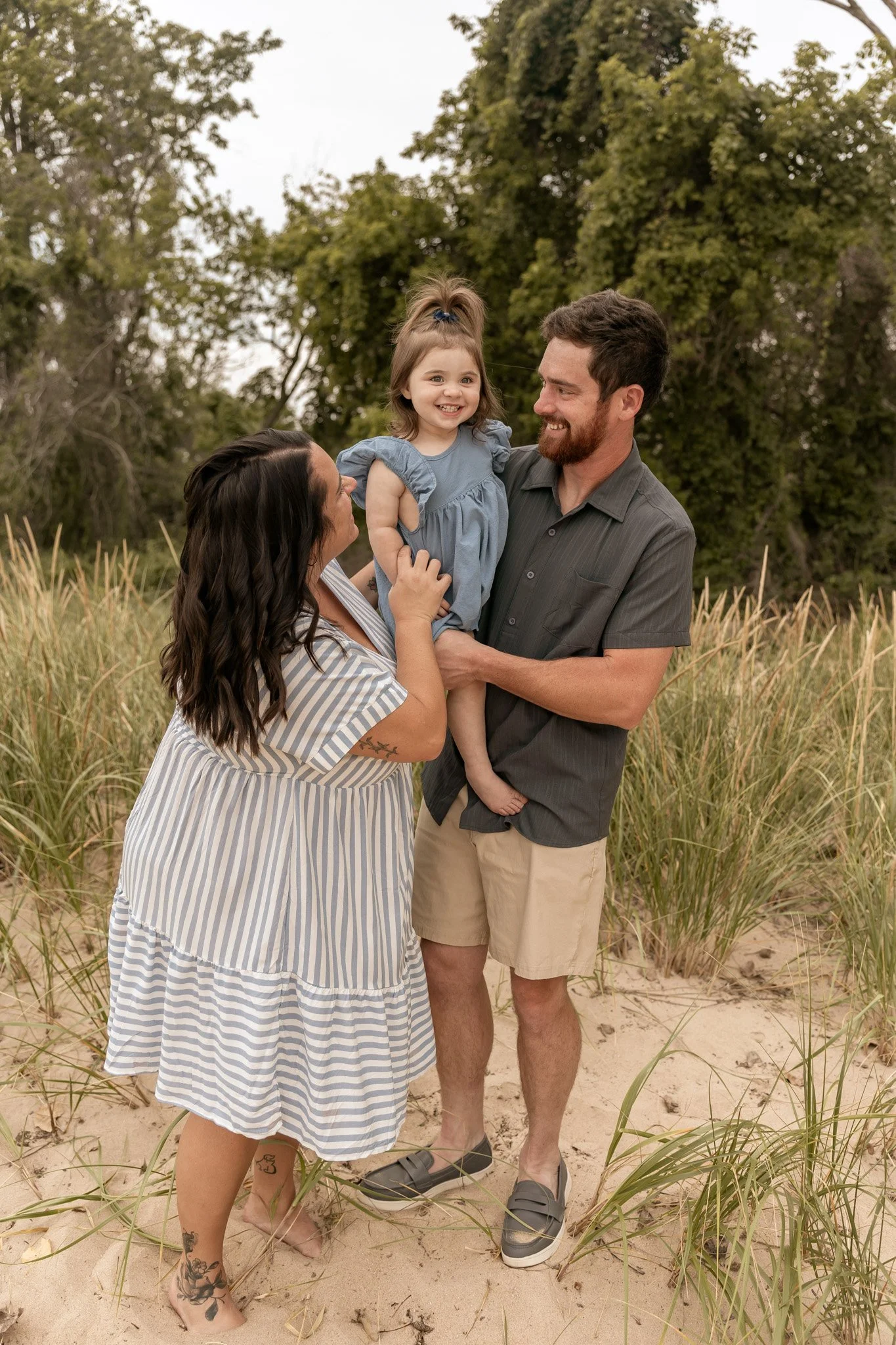 A family of three standing on a beach with tall grass. The dad is holding a smiling young girl, and the mother is touching her face. Photo by Feinauer Photography in Bay City, Michigan.