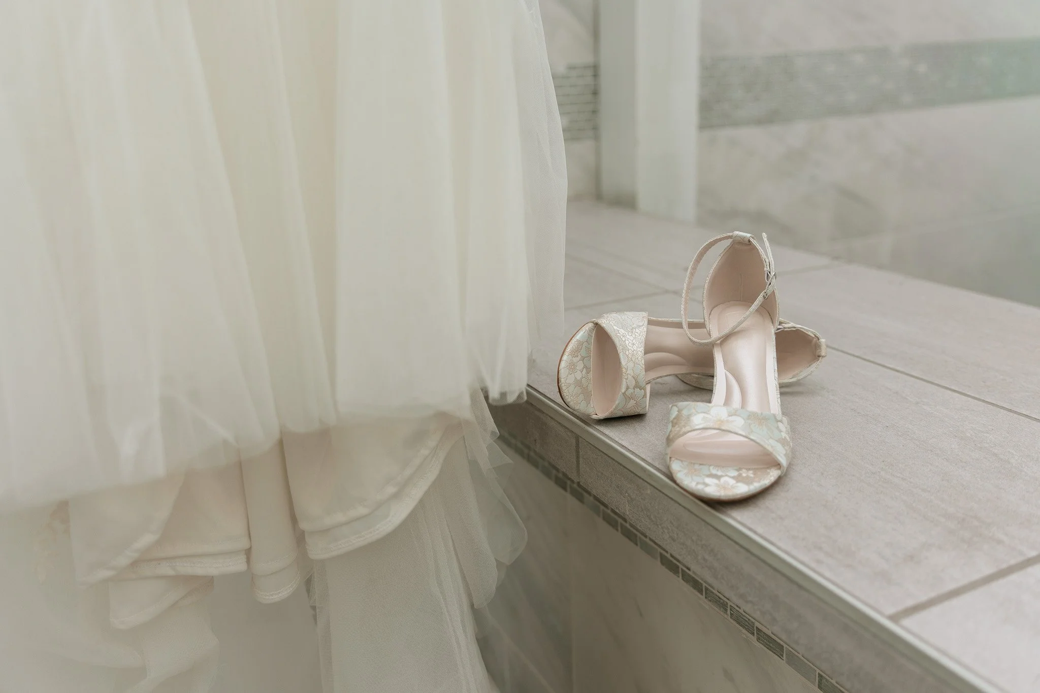 A pair of beige floral high-heeled shoes with a touch of sometime blue on an outdoor bar next to wedding dress