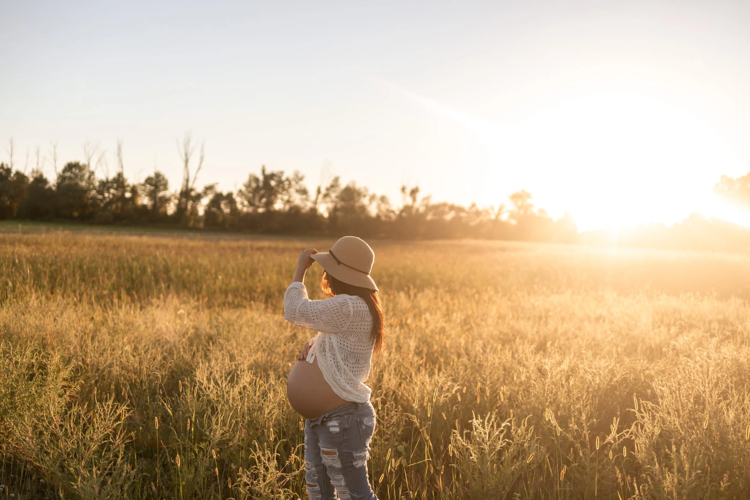A pregnant woman wearing a white crochet top, ripped jeans, and a wide-brimmed hat standing in a field at sunset, with trees in the background.