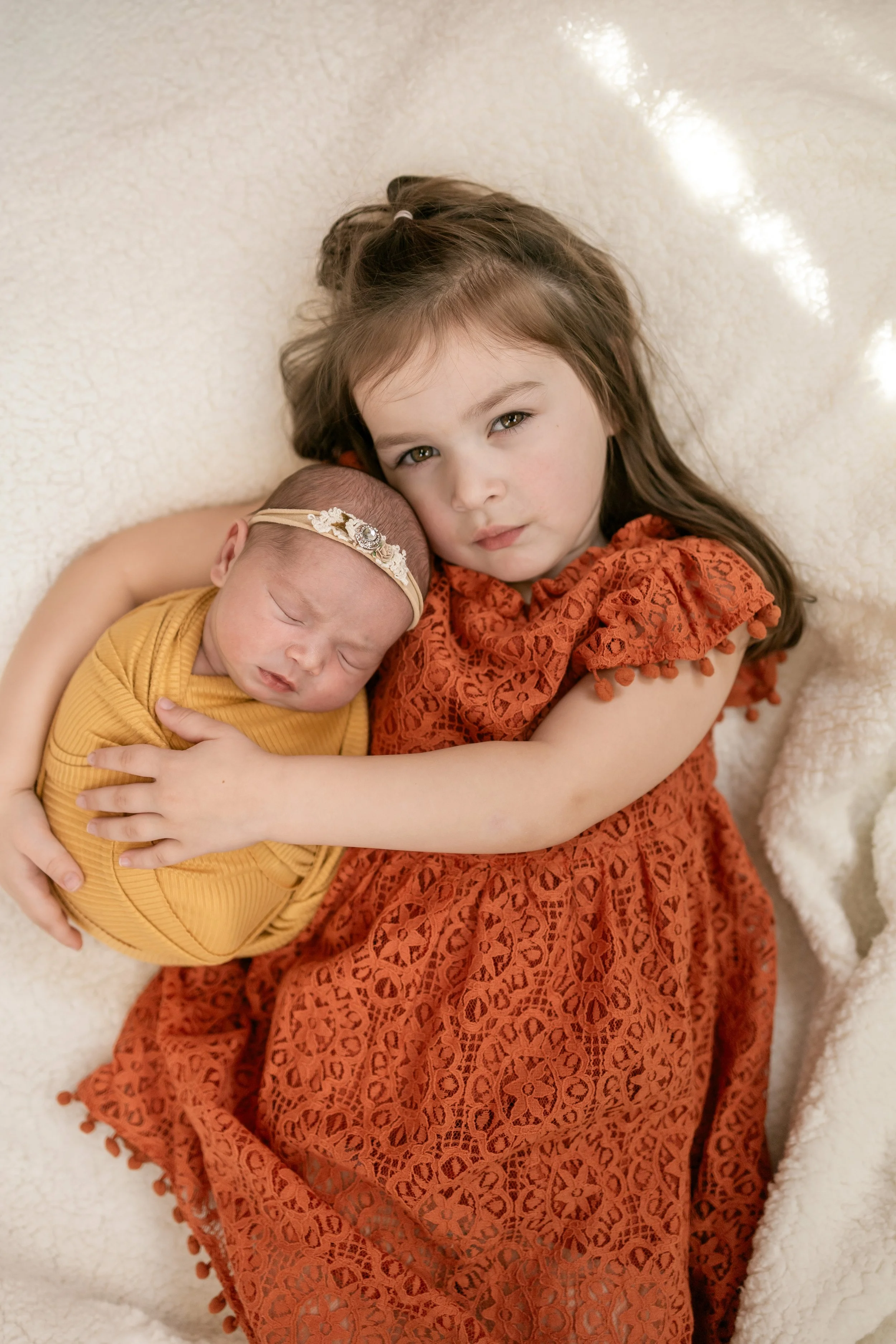 A young girl in an orange lace dress holds a sleeping newborn girl in a yellow dress, both lying on a soft cream-colored blanket.