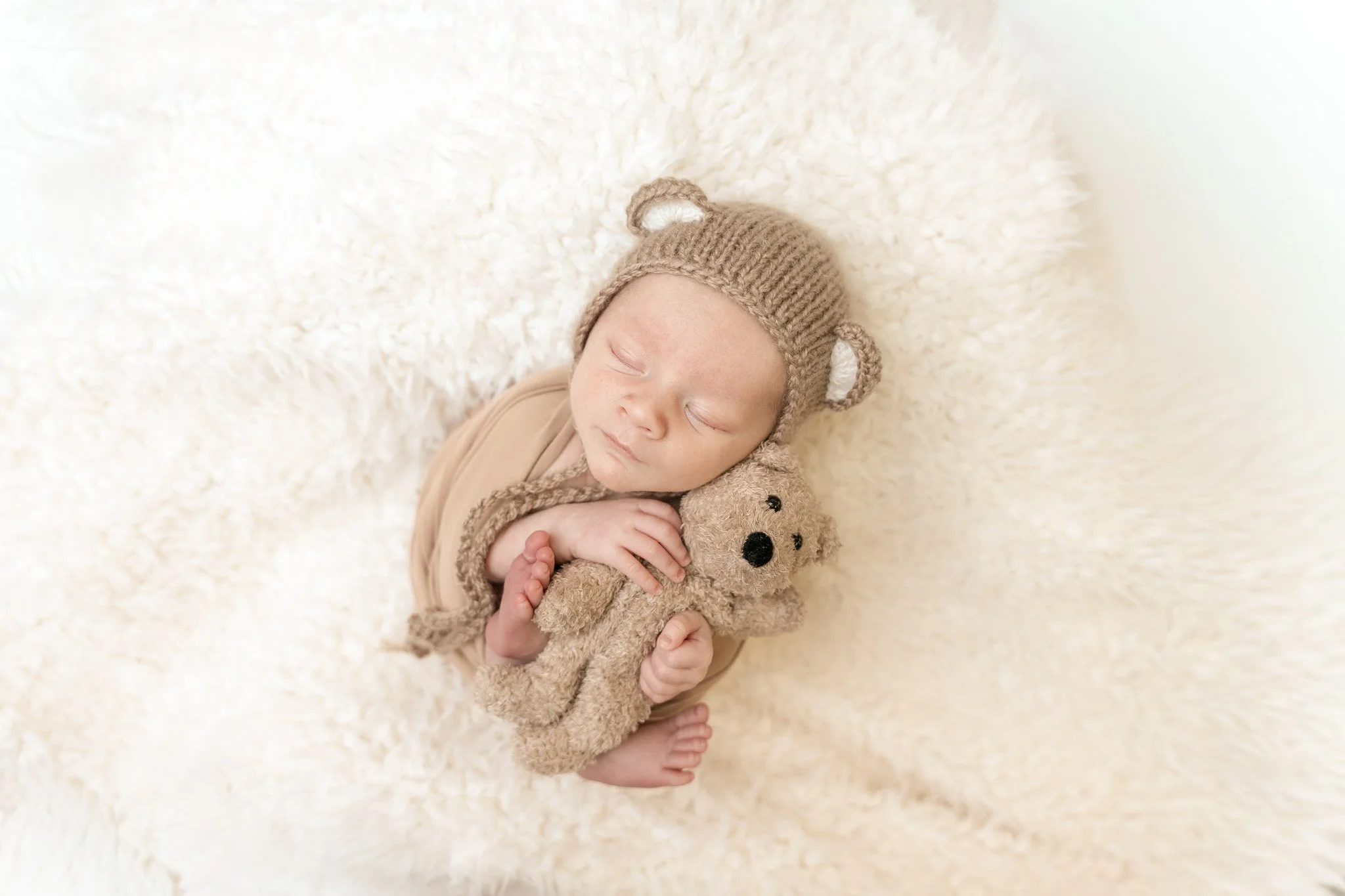 Sleeping newborn baby wearing a knitted bear hat and cuddling a stuffed teddy bear on a soft cream blanket