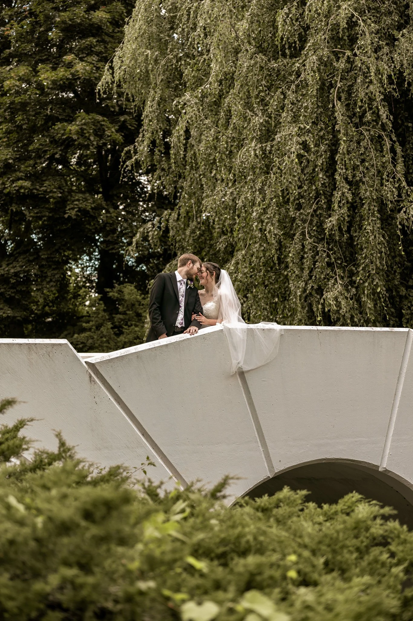 A bride and groom hugging on a white bridge with lush green trees in the background.