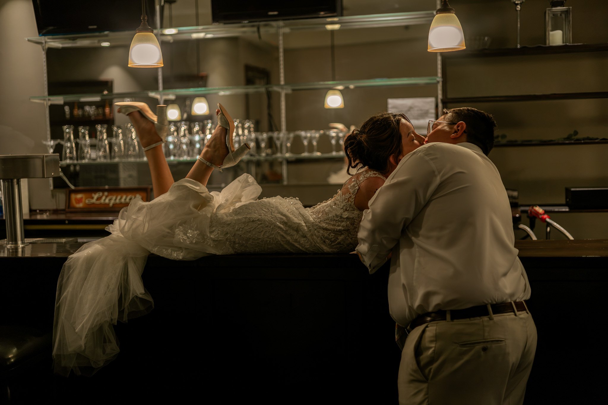 A bride and groom share a kiss at a bar, with the bride lying on the counter and the groom kneeling in front of her, in a dimly lit setting.
