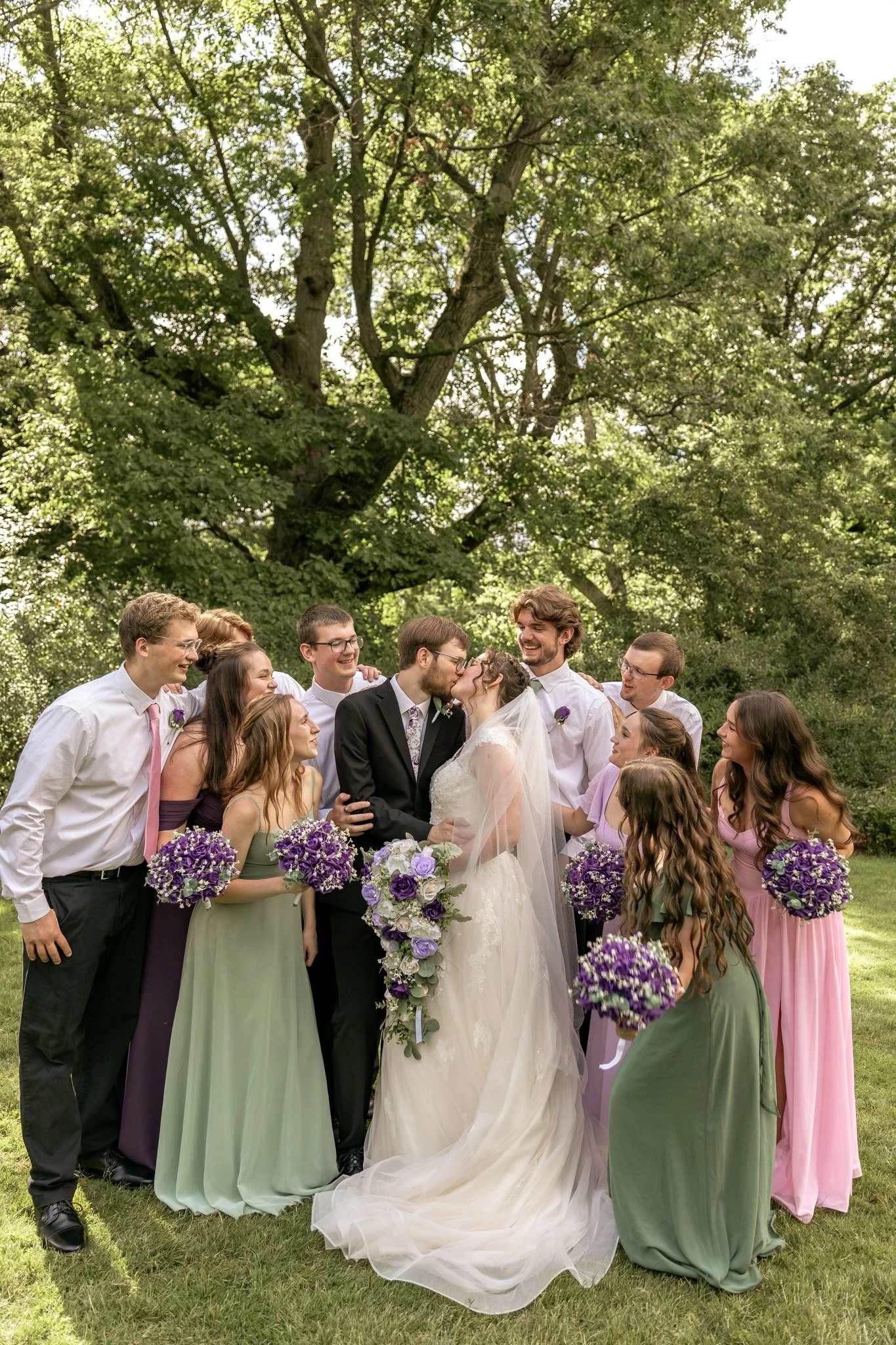 A group of people gathered outdoors under a large leafy tree, celebrating a wedding. The bride and groom are in the center, kissing, with the bride in a white wedding dress holding a bouquet of purple and white flowers. The bridesmaids wear pastel-co