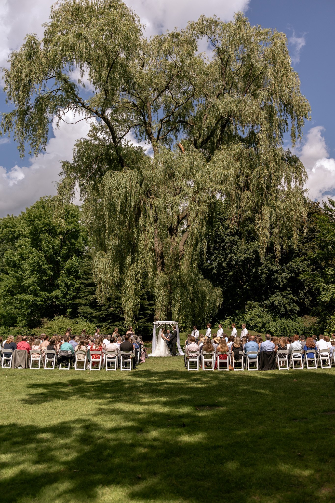 An outdoor wedding ceremony taking place in a lush green park under a large leafy tree. There is a bride and groom exchanging vows at an arch, surrounded by guests seated on white chairs.