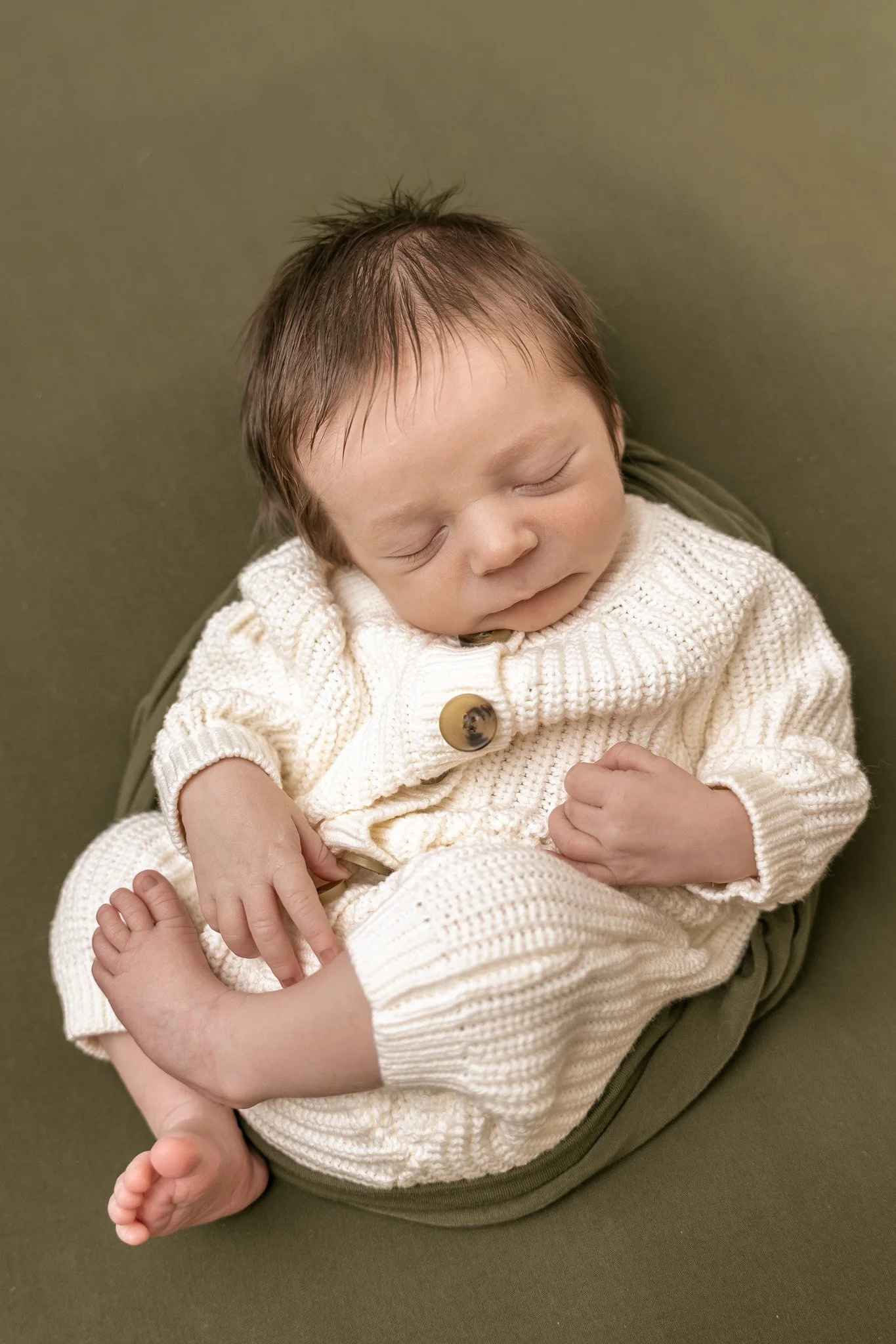Sleeping baby wearing a cream knitted sweater and sitting on an olive-green blanket