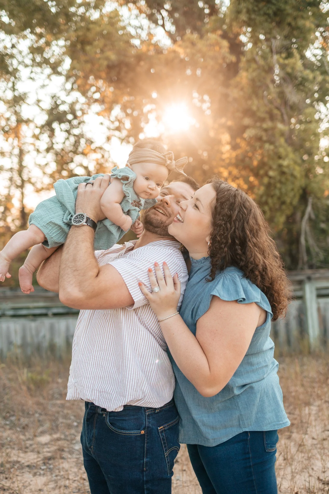 Family of three, a man, woman, and baby girl, smiling and embracing outdoors with trees and sunlight in the background.