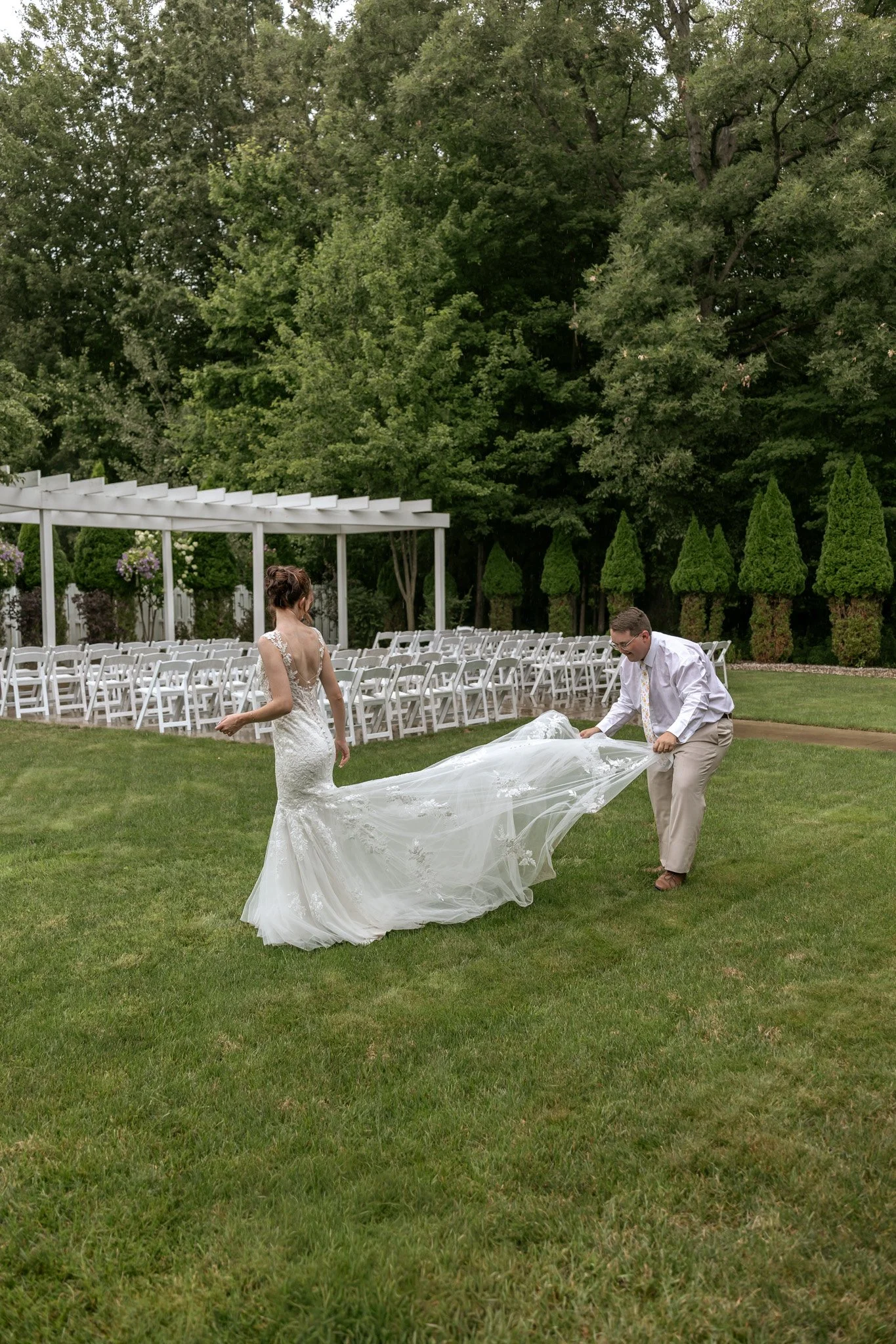 A bride and groom preparing for a wedding outdoors on a grassy area with rows of white chairs and lush green trees in the background.