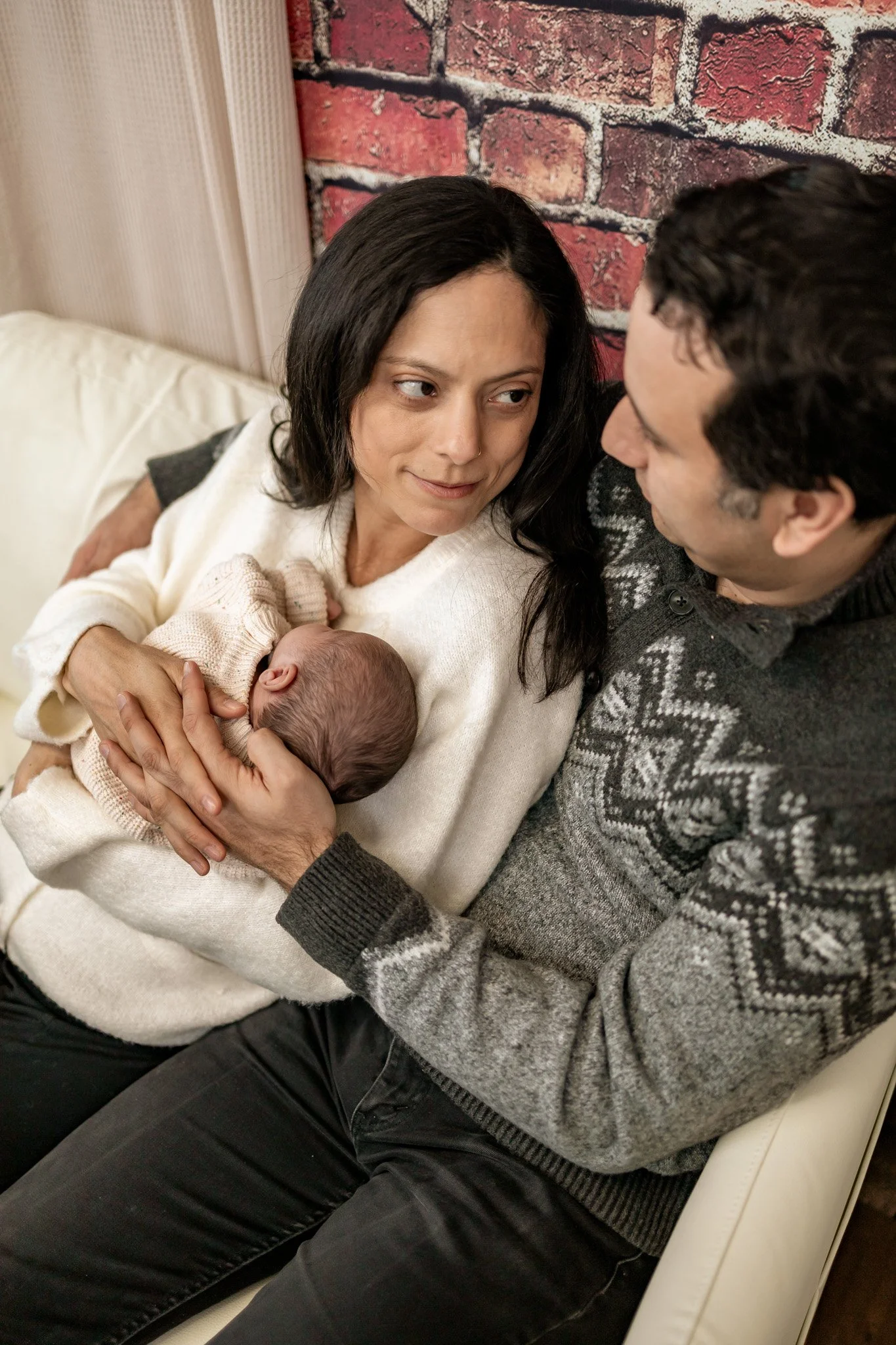 A woman holding a newborn baby, with her head tilted towards a man sitting nearby, in a cozy indoor setting with a brick wall background.