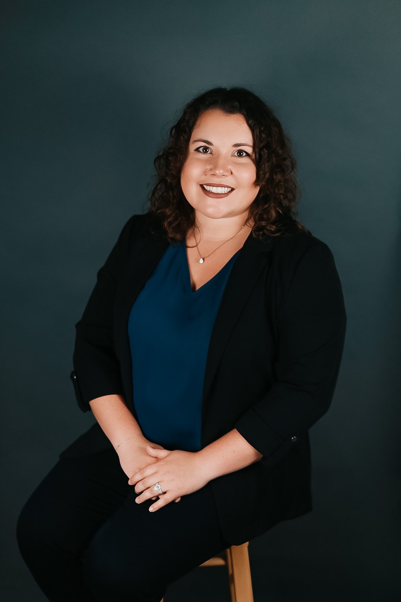 Portrait of a woman with curly hair, wearing a blue top, black blazer, and black pants, sitting on a stool against a dark background.