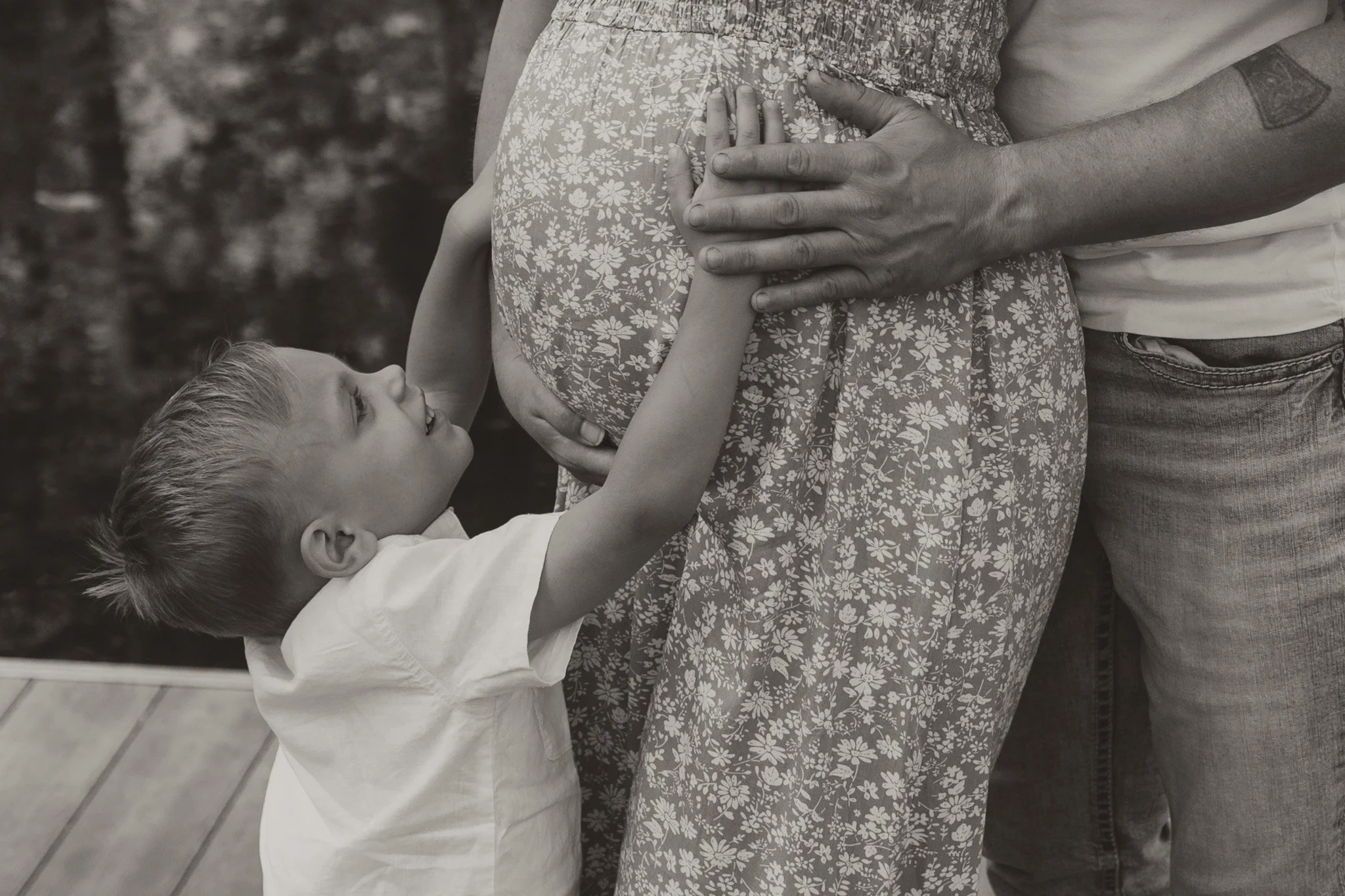 A young boy reaches up and touches a pregnant woman's belly, with a man's hand resting on her stomach, in an affectionate moment.