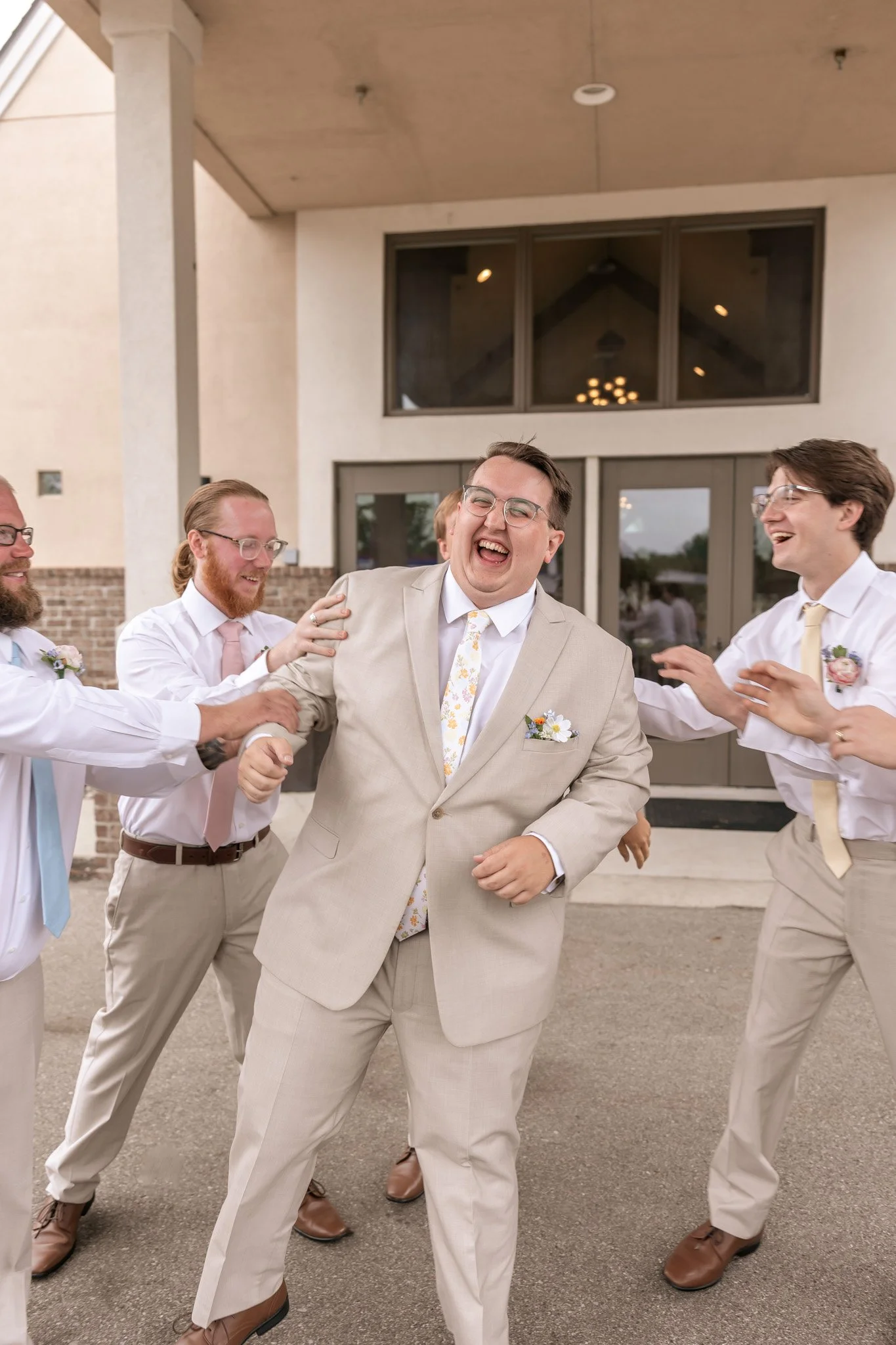 Groomsmen dressed in beige suits and white shirts are gathered outside a building, laughing and celebrating. They are holding hands and embracing each other, indicating a joyful occasion.