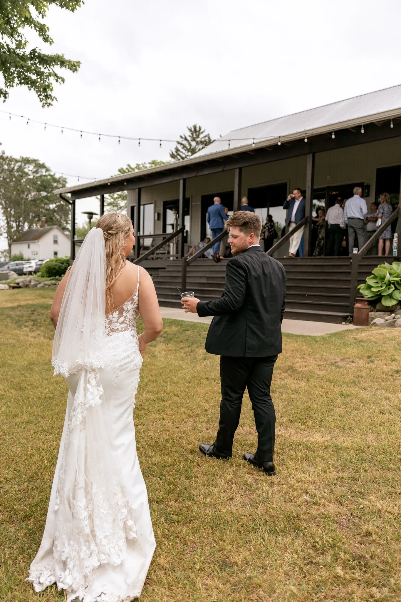 Bride and groom standing outdoors on grass at a wedding reception, with guests socializing on a wooden deck in the background and string lights hanging overhead.