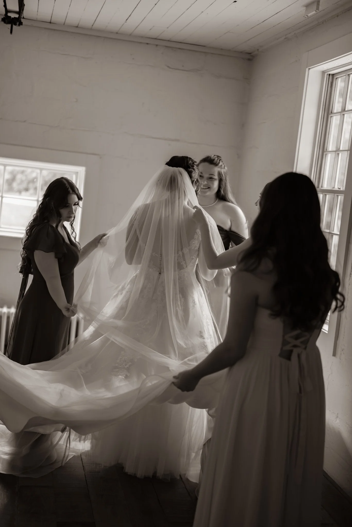 Women helping bride put on wedding dress in a bright room with large windows.