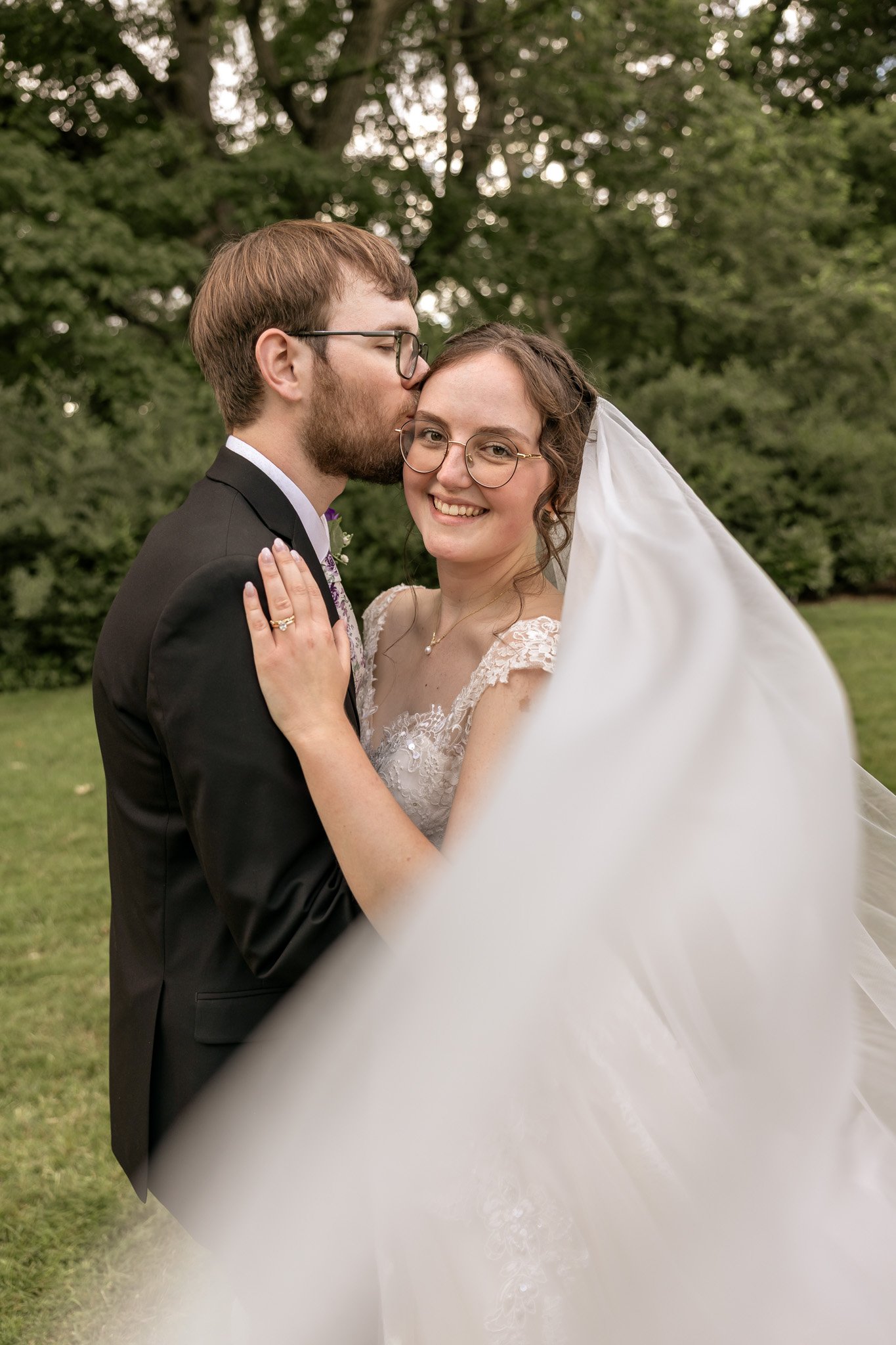 A newlywed couple standing outdoors with greenery in the background; the groom, with glasses and a beard, is kissing the bride on her temple, who is smiling and wearing glasses and a wedding dress with a veil.