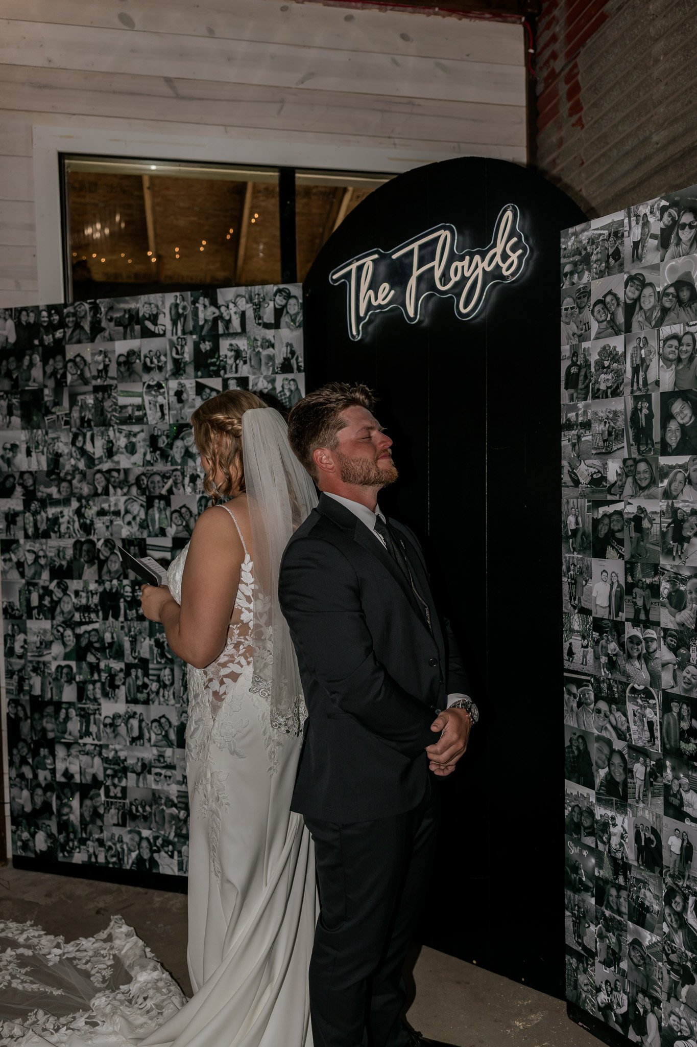 A groom with his eyes closed standing in front of a photo display at a wedding, with a bride in a wedding dress behind him, both facing away from each other.