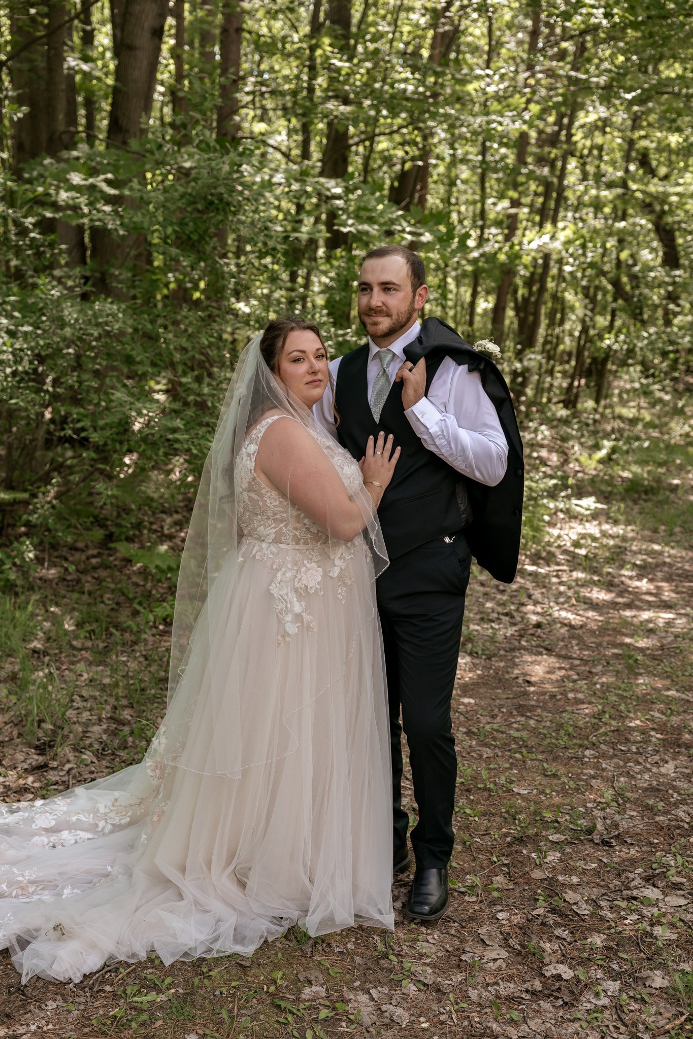 A bride and groom standing together in a wooded area. The bride is wearing a white lace wedding dress and veil, and the groom is wearing a black vest, white shirt, and tie, holding a black jacket over his shoulder.