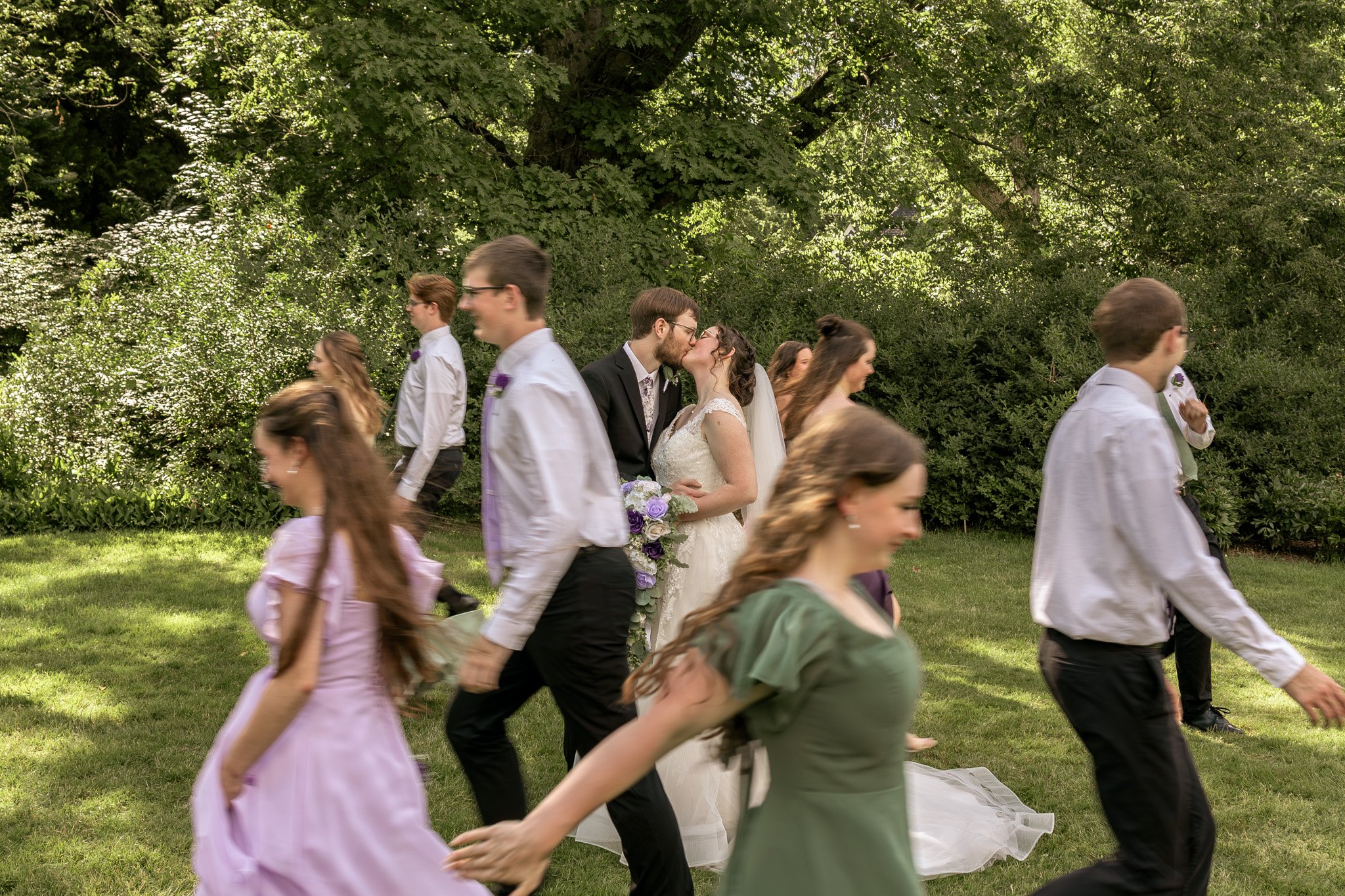 A group of people, including a bride and groom, holding hands and walking outdoors in a park with green trees. The bride is wearing a white wedding dress, and the groom is in a black suit. Others are dressed casually, and some people are blurred as t