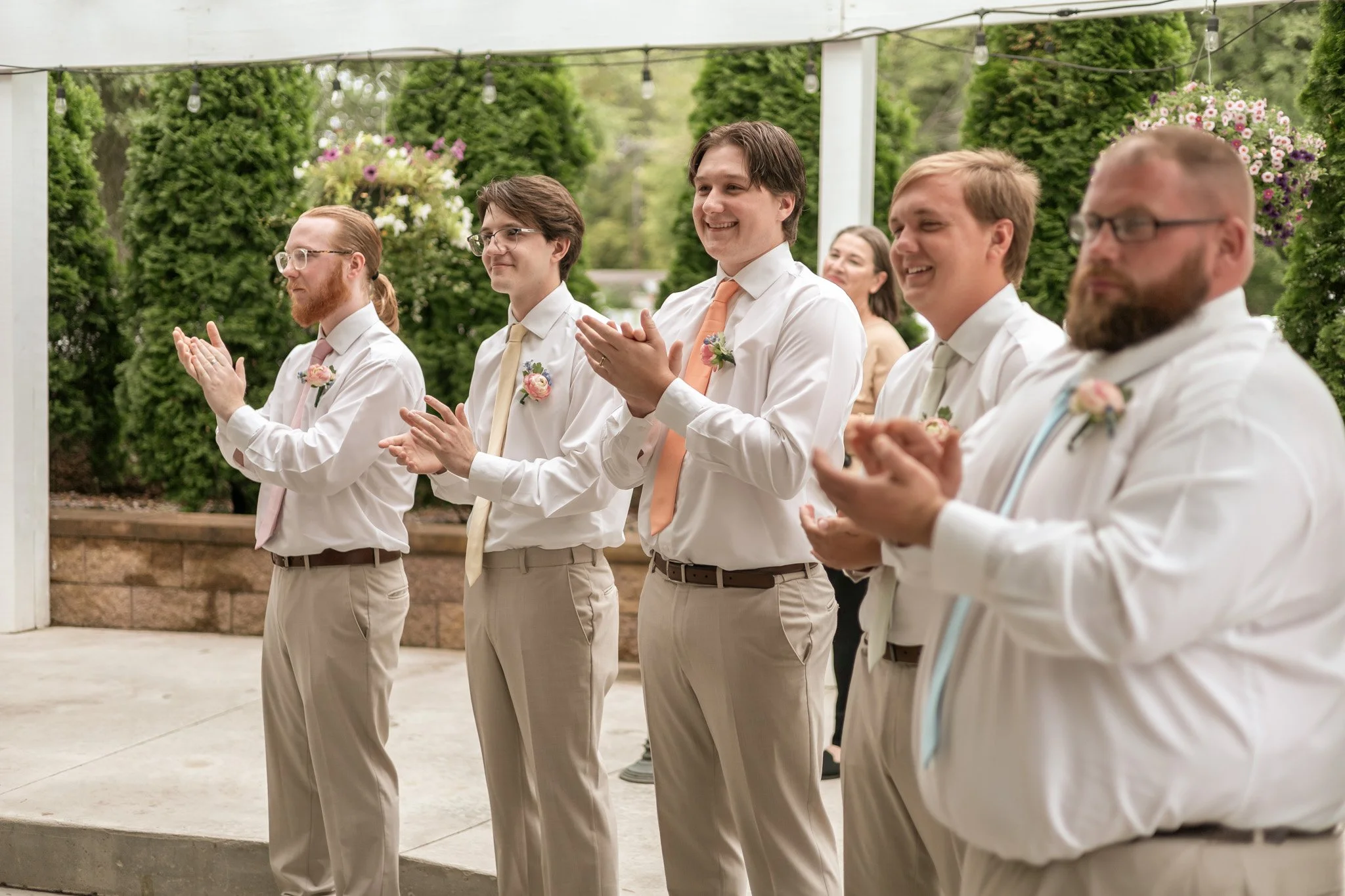 Groomsmens dressed in white shirts and beige pants standing in a line, clapping, and smiling at an outdoor event with greenery and hanging flowers in the background.