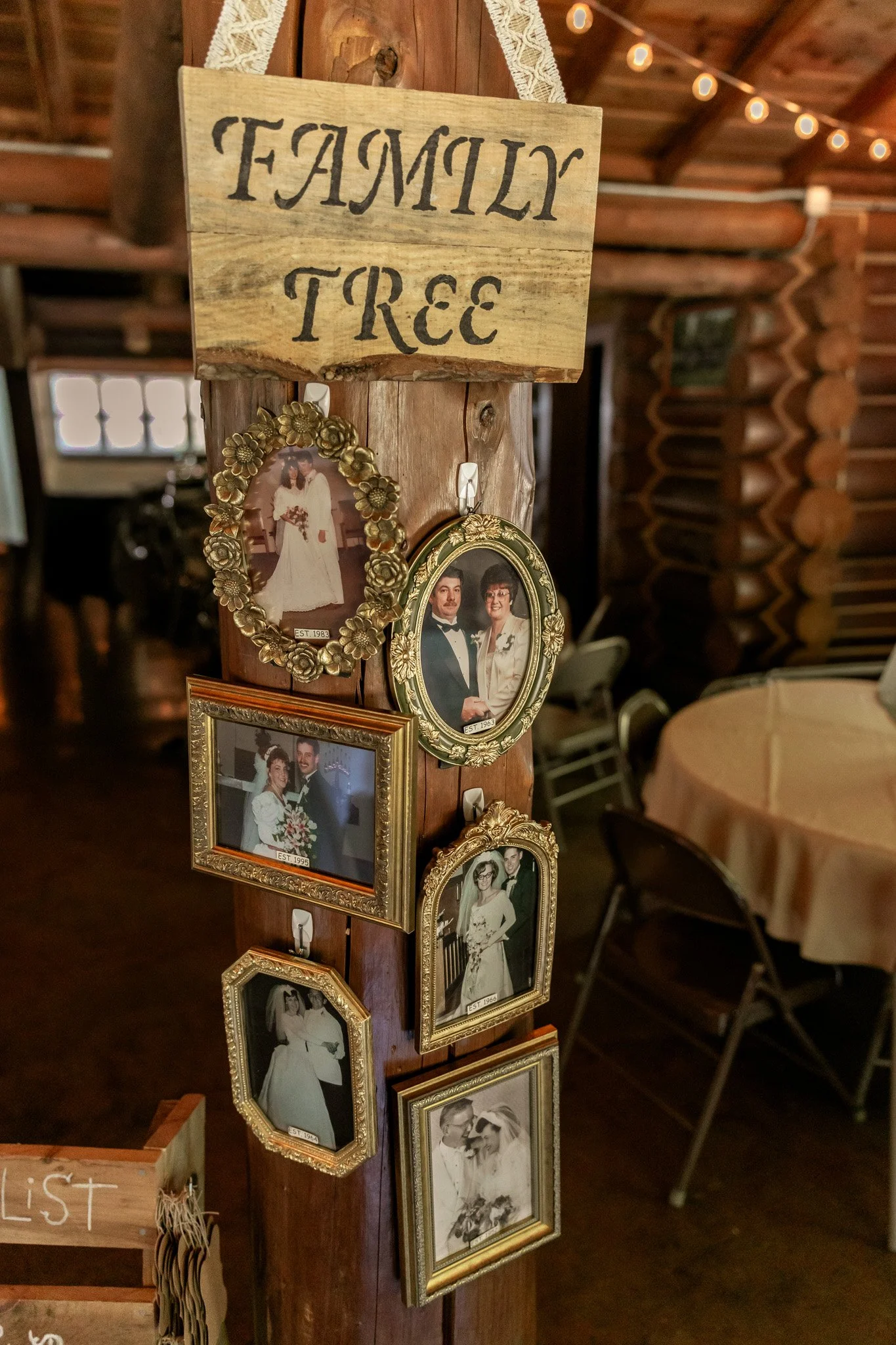 A wooden sign with the words 'FAMILY TREE' painted on it hangs on a wooden post. Below the sign are several framed vintage-style photographs of couples, likely family members, arranged along the post. The background shows a cozy rustic interior with 