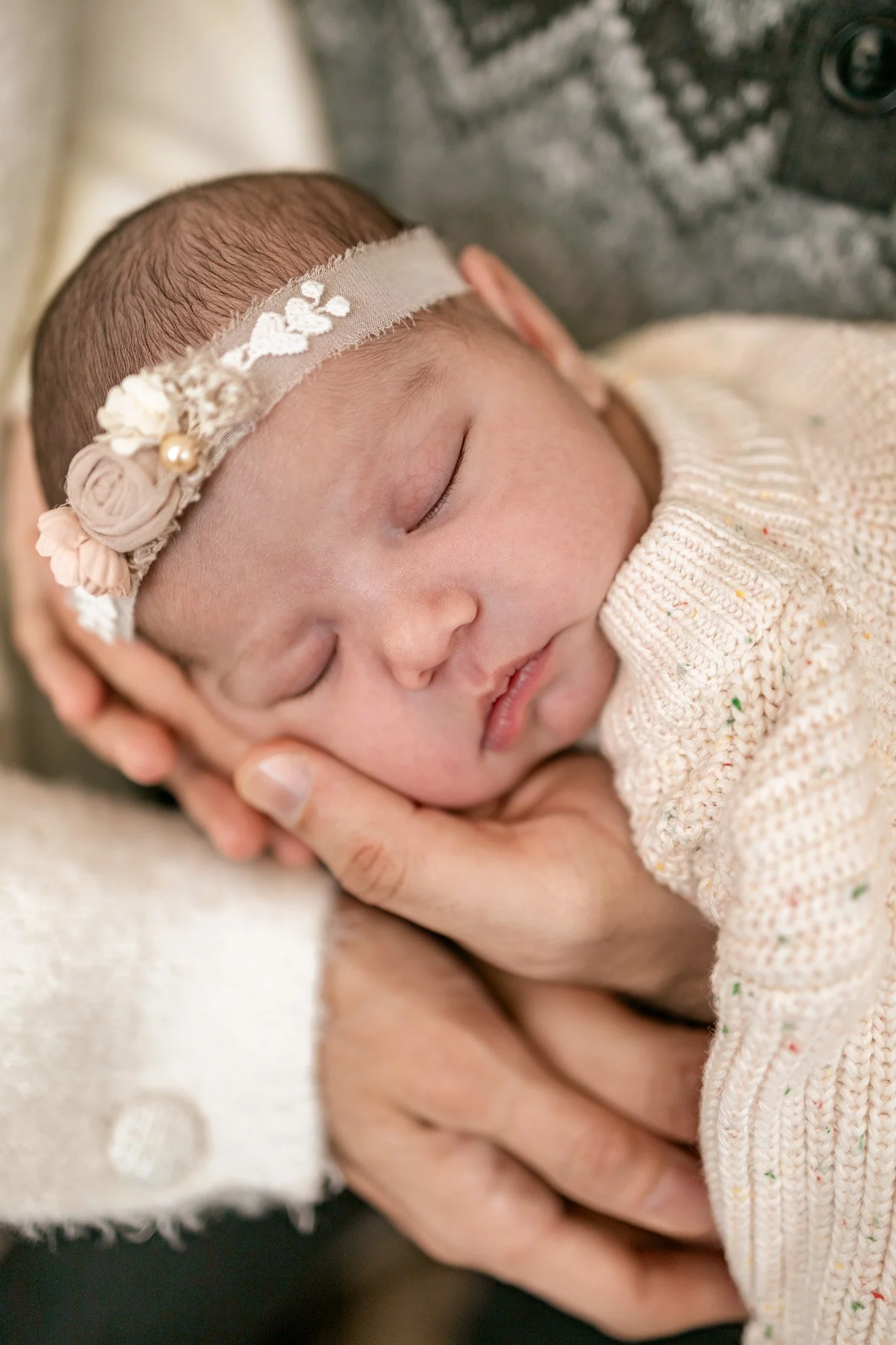 Close-up of a sleeping newborn baby with a floral headband, cradled in someone’s hand.