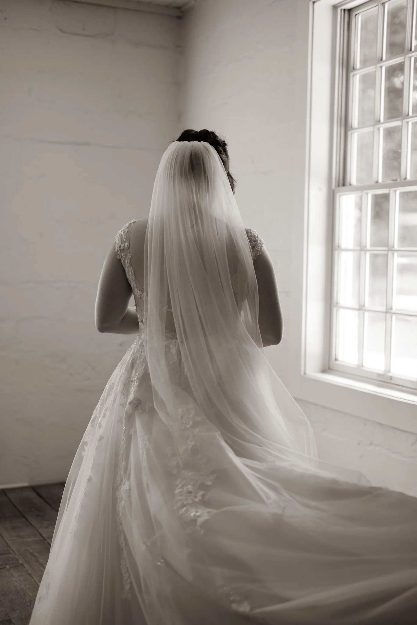 Bride in wedding dress with veil, standing by window in a simple, bright room.