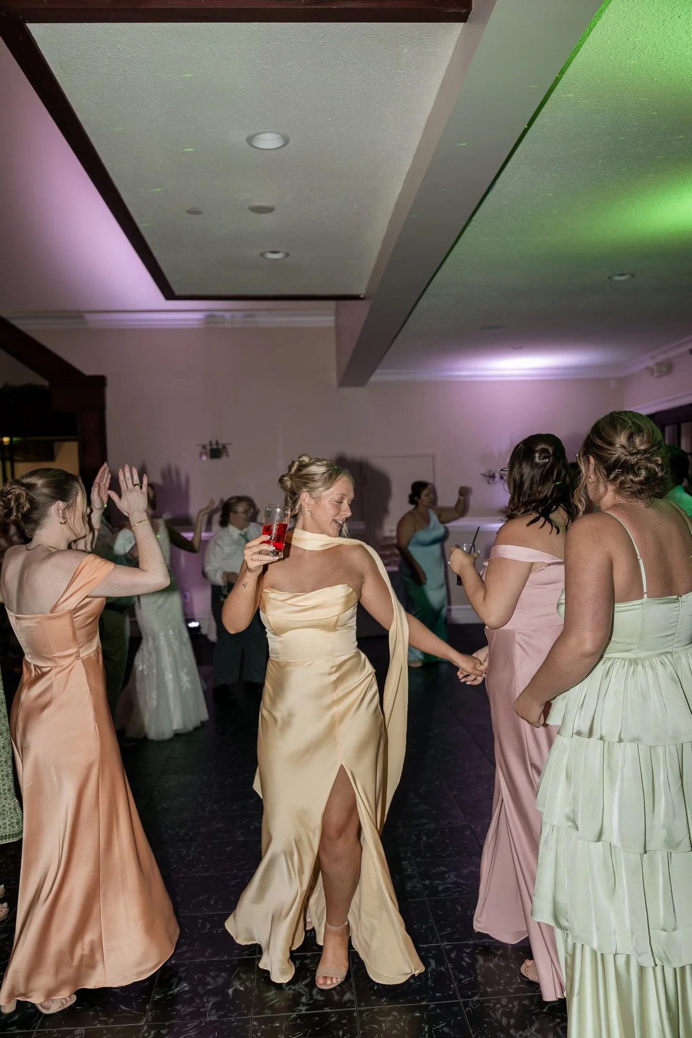 A group of women dancing and enjoying themselves at a party, wearing colorful satin dresses in shades of peach, yellow, pink, and green.